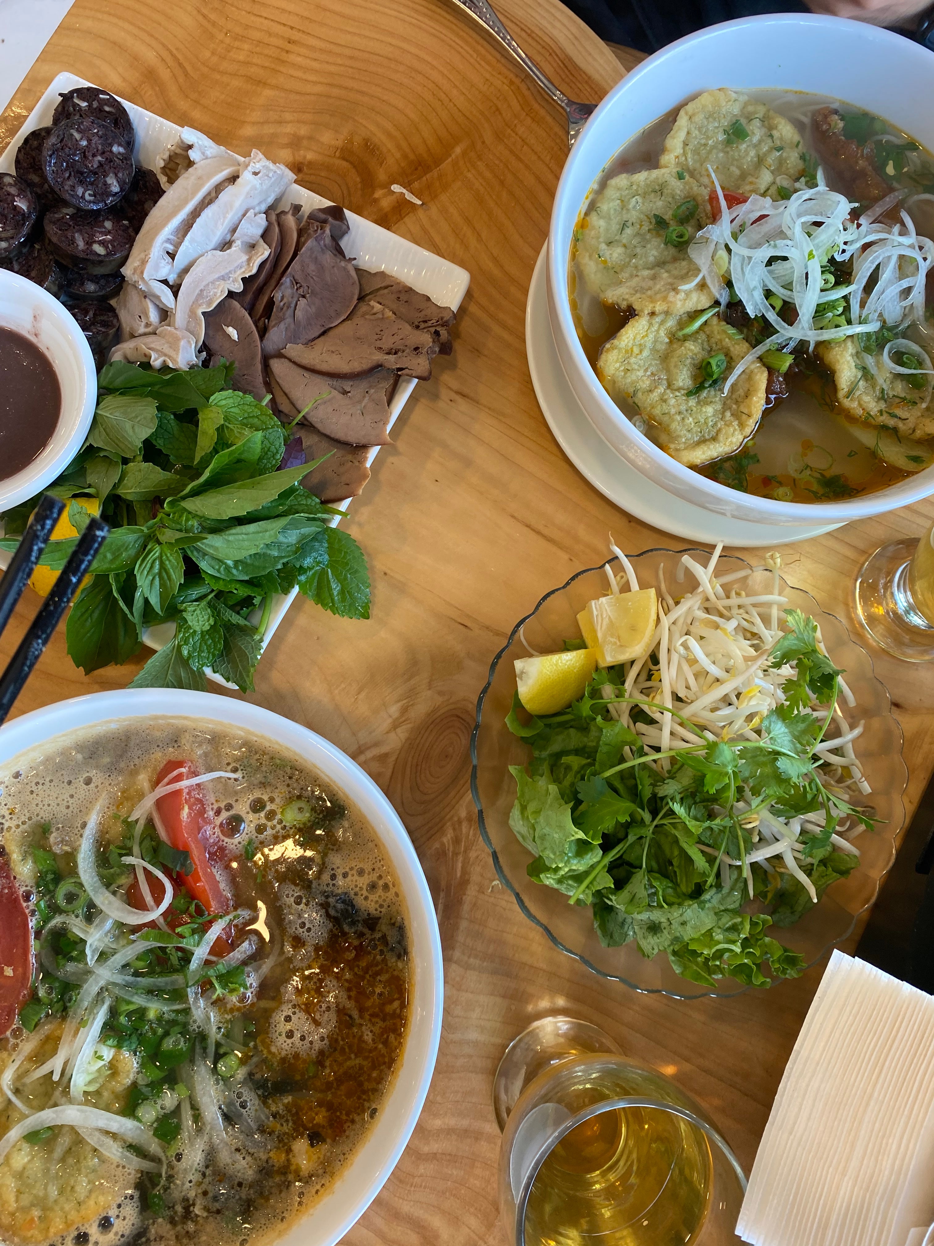 An assortment of food is on a table including vegetables, meats, noodle soups and drinks. 