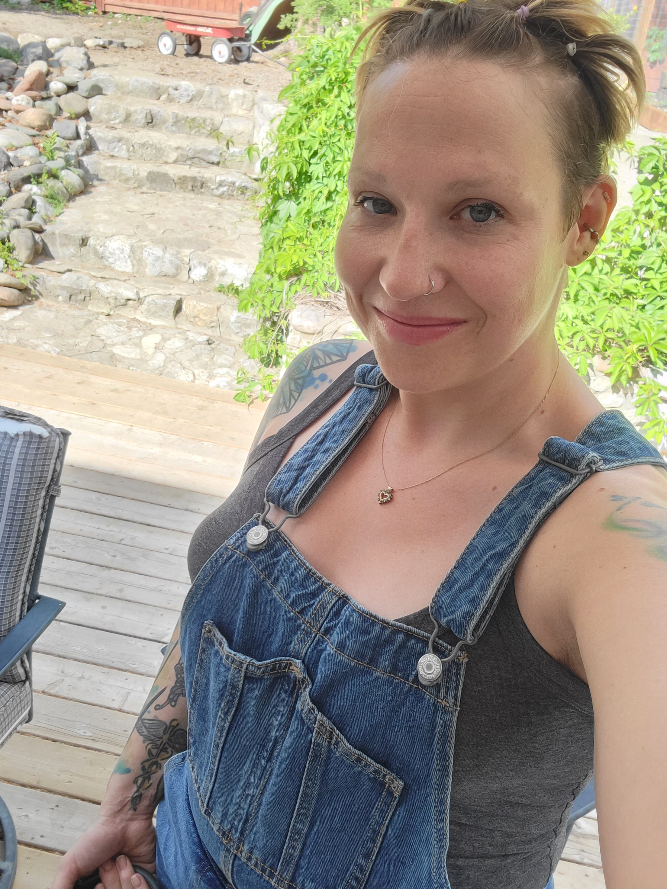 A woman with short spiky brown hair, nose ring and tattoos on her shoulders wearing jeans overalls takes a selfie in front of stone stairs and a wooden deck