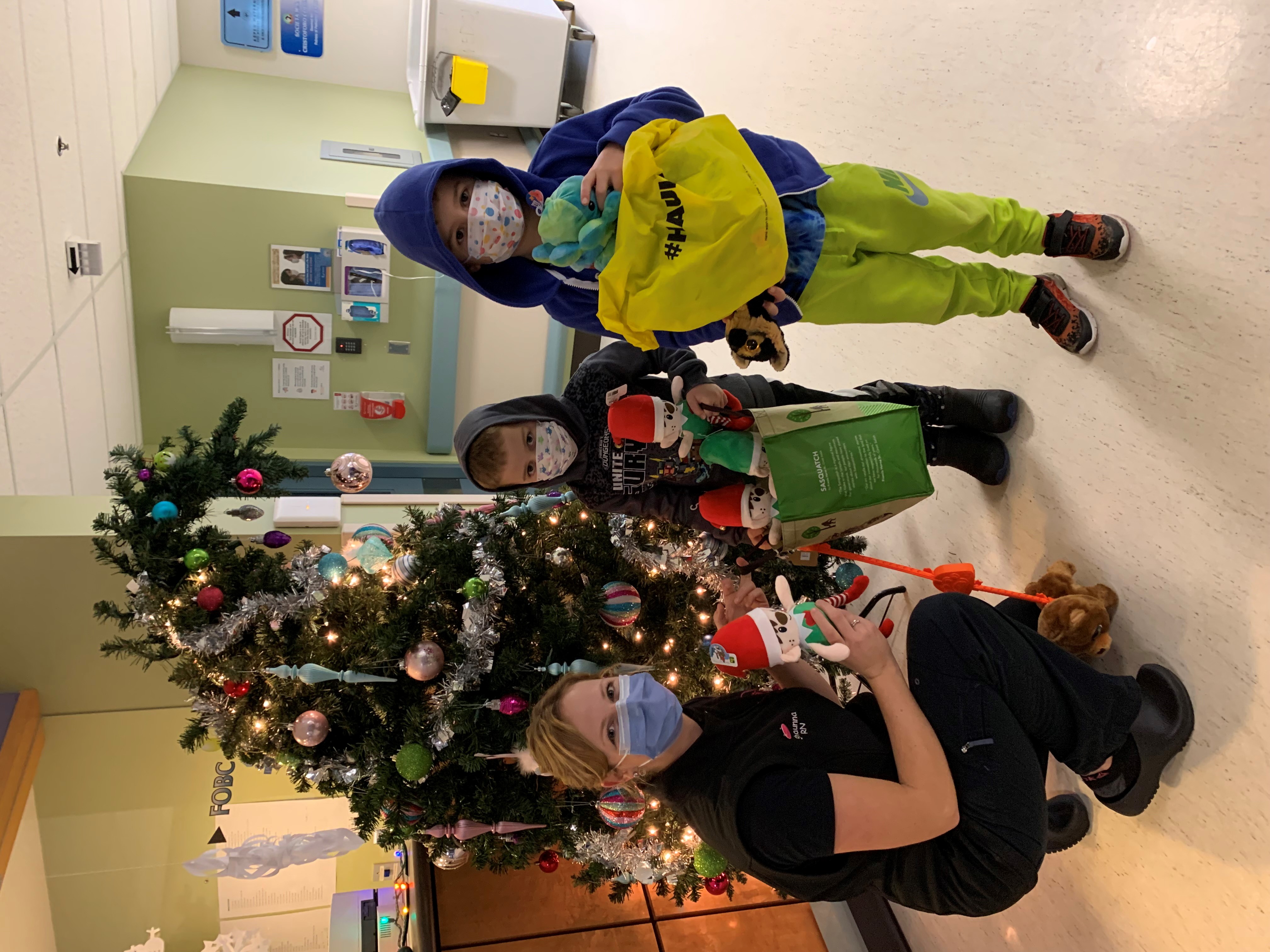 A health-care professional and two youths holds presents near a Christmas tree in a hospital area.