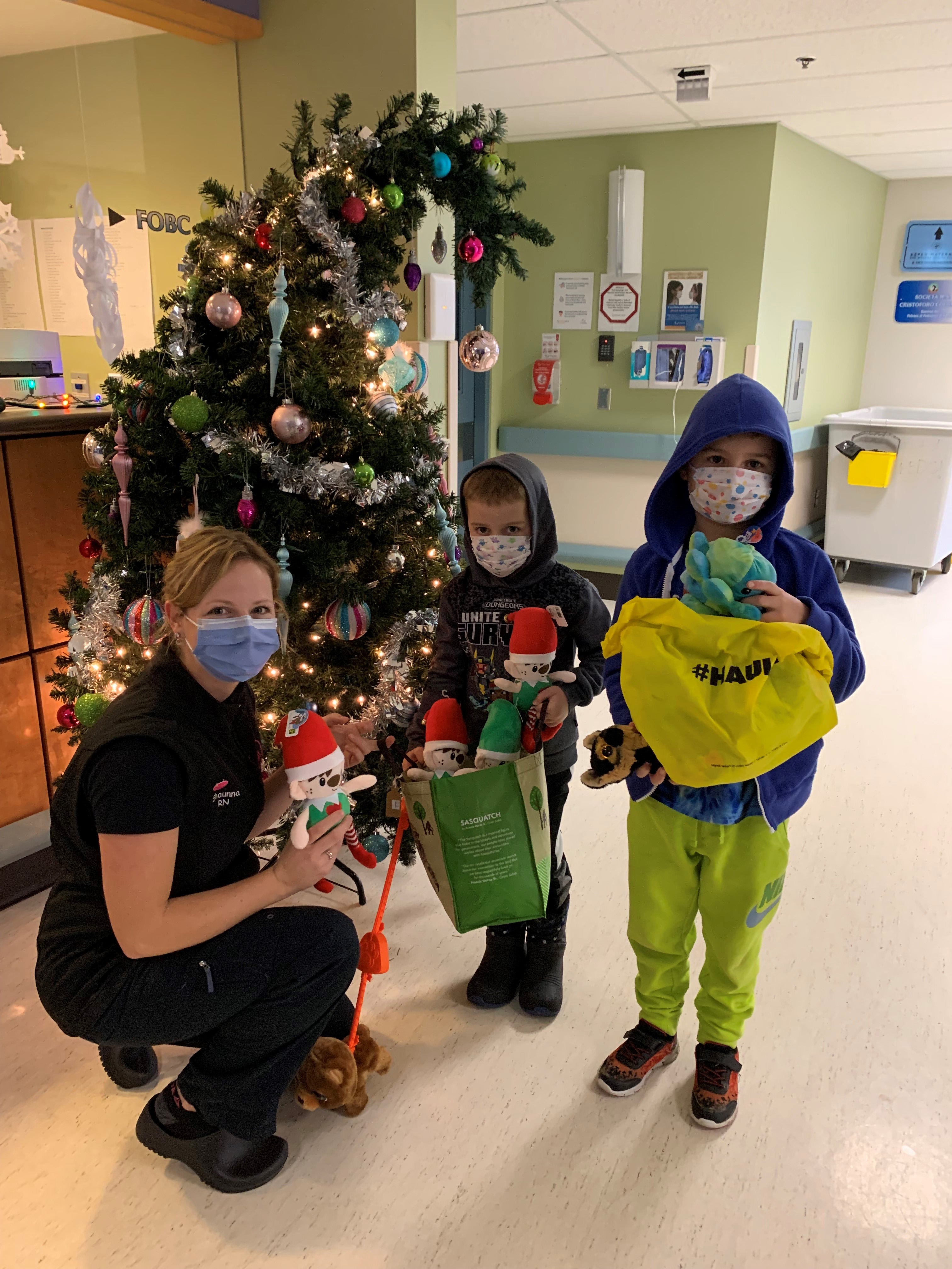 A health-care professional and two youths holds presents near a Christmas tree in a hospital area.