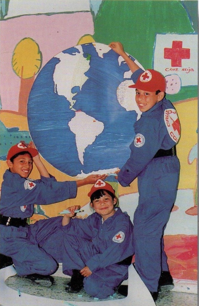 Three children in Red Cross uniforms pose around a painted globe mural.
