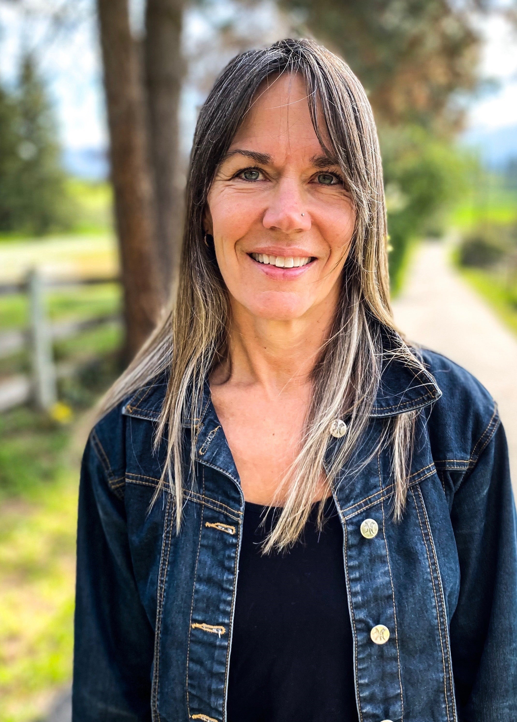 A person with long grey-blonde hair with bangs wearing a jean jacket stands in front of a country lane with trees and a fence.