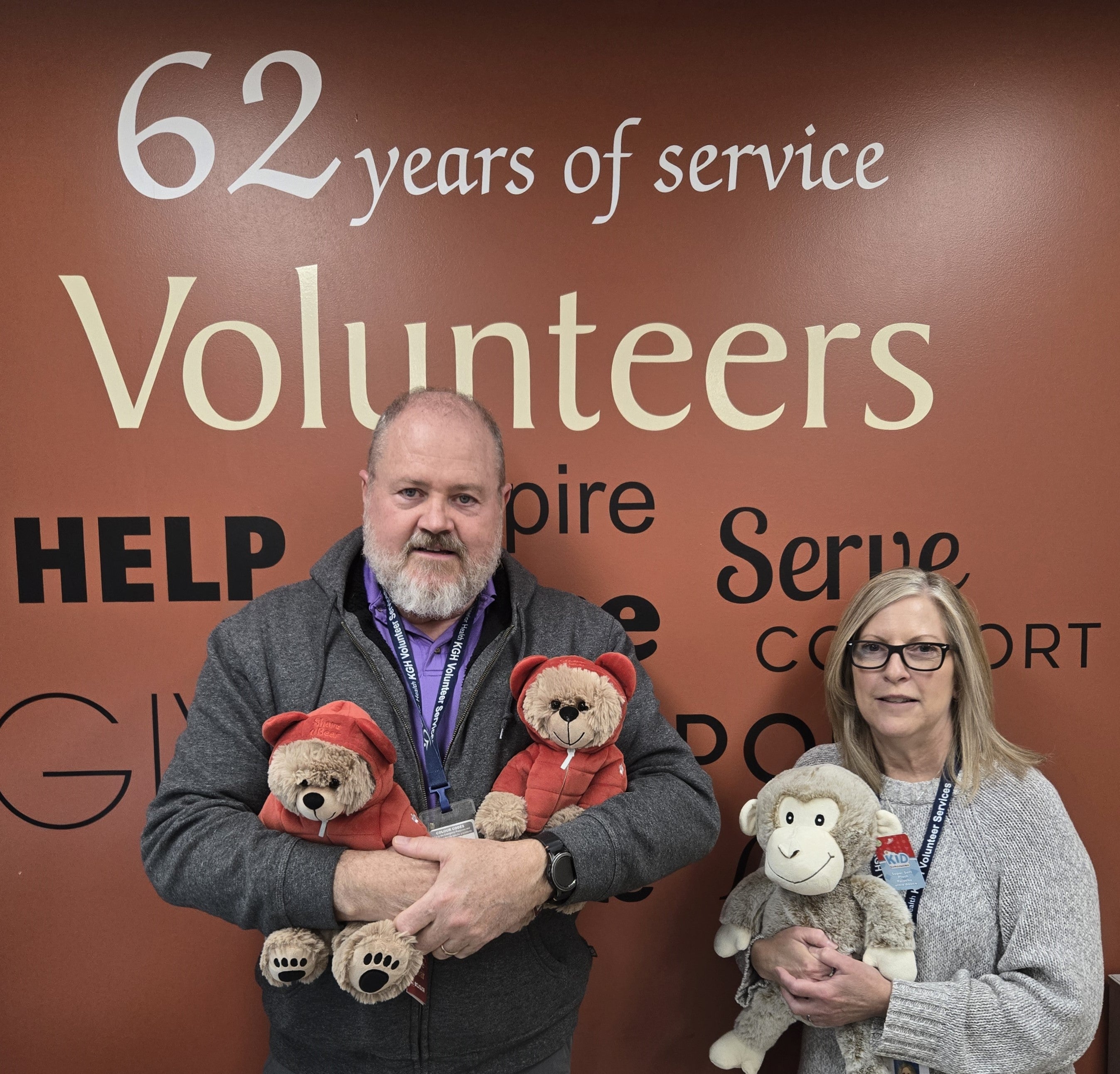 Two people holding stuffed toys in front of a wall with text, including the words 62 years of service and the word volunteers.