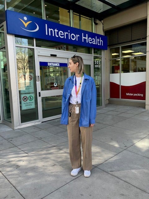 Woman with shoulder-length hair, wearing a white t-shirt, blue overshirt, and brown pants, stands in front of a building that reads "Interior Health"