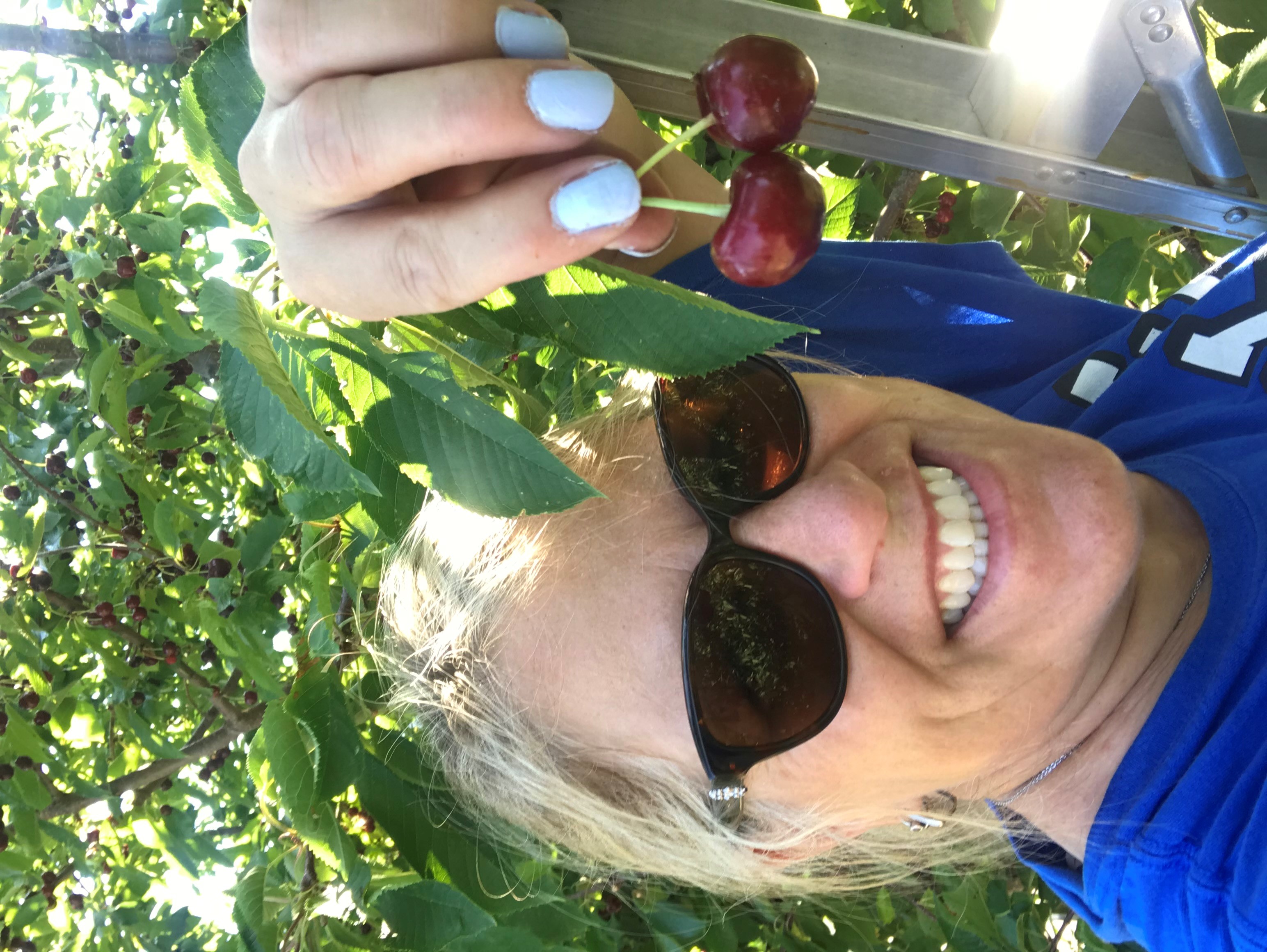 A woman with sunglasses on taking a selfie in her garden while holding up freshly picked cherries