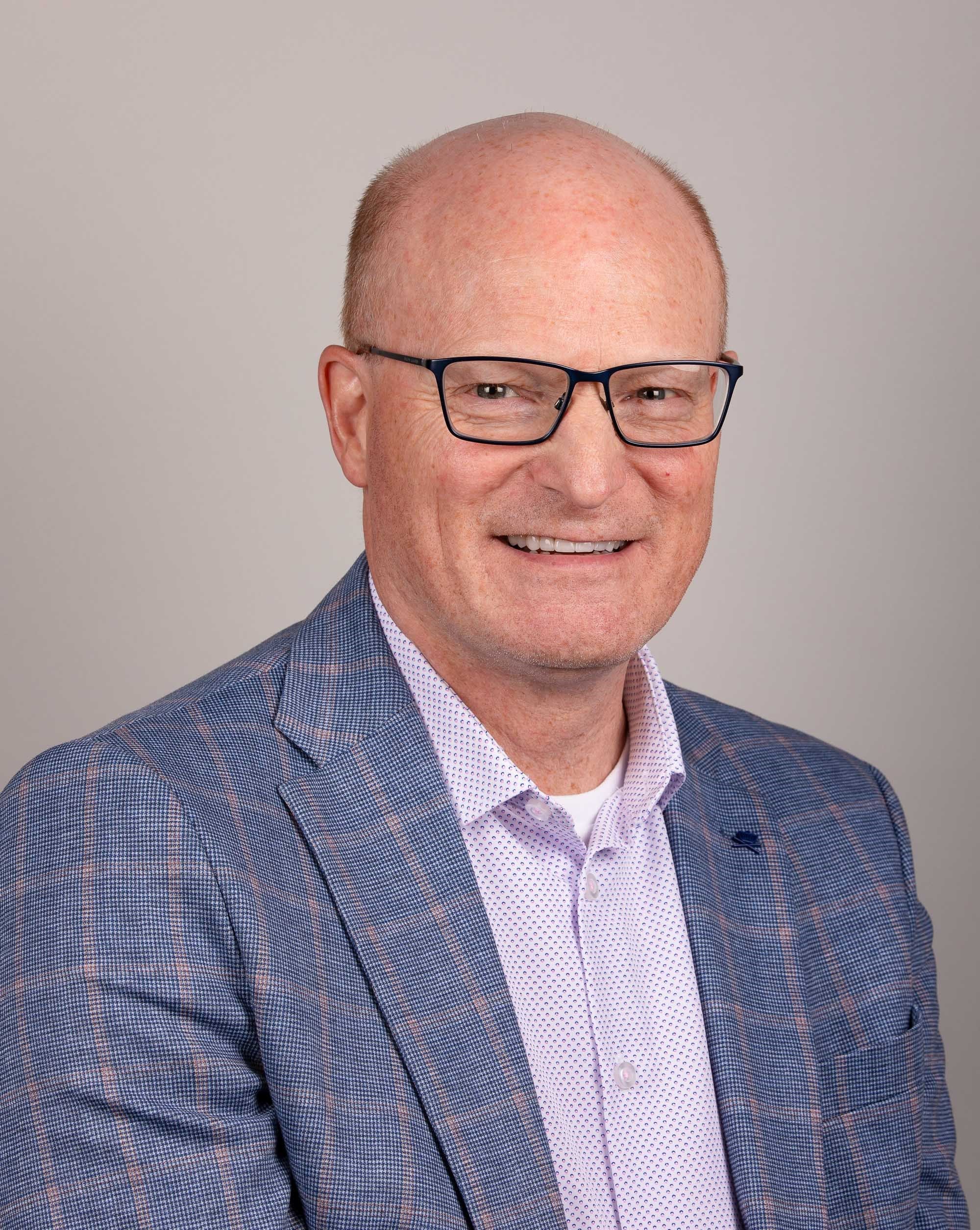 person with glasses and very short hair smiles at the camera while wearing suit