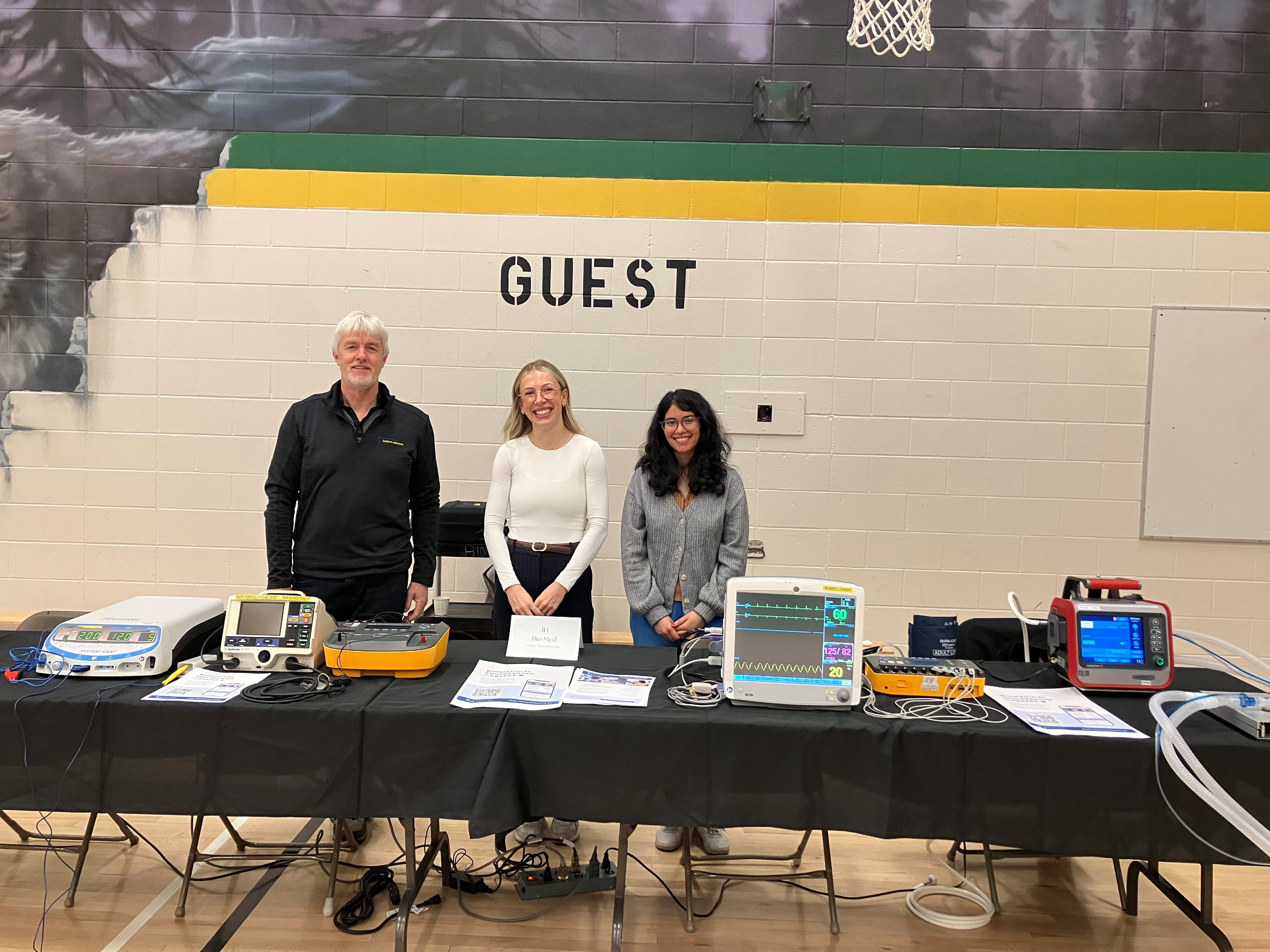 Three people stand behind a table showing various kinds of monitors and biomedical equipment used in medical settings.