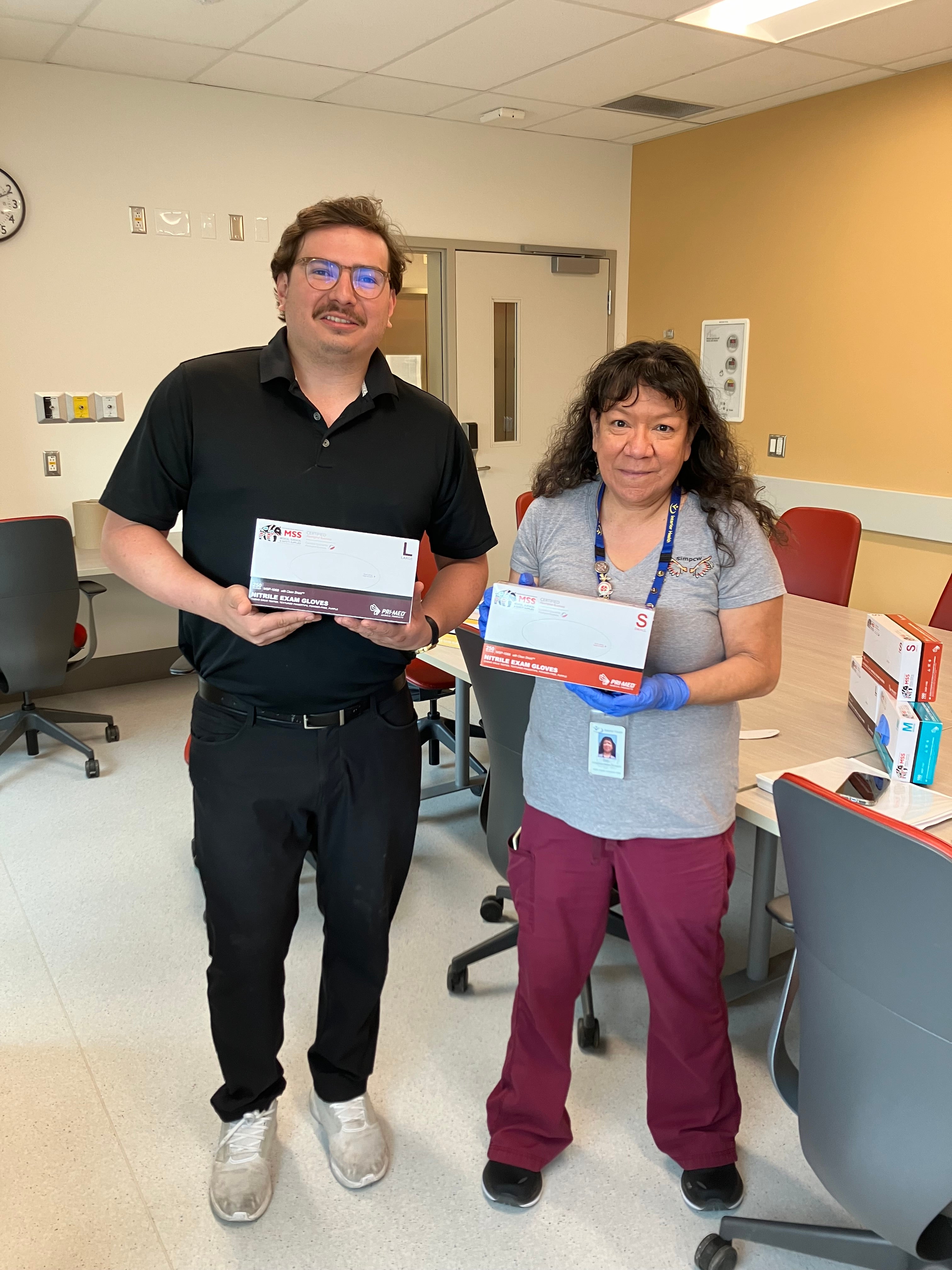 Man wearing all black stands next to a hospital staff member wearing scrubs, both are holding a box of gloves.