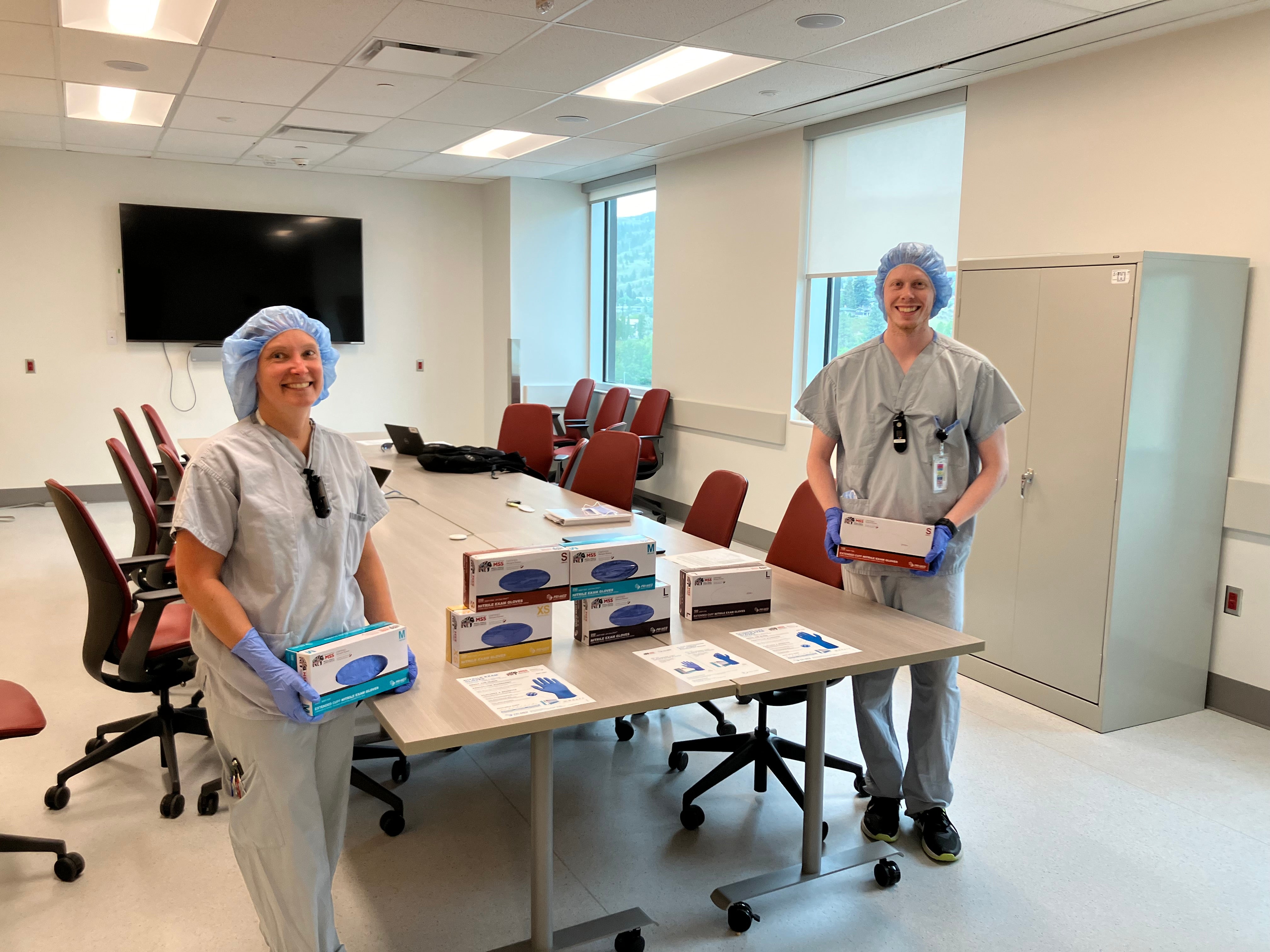 Two medical staff wearing scrubs, scrub caps, gloves, and one stethoscope around their neck are holding a box of gloves standing on either side of a table with a boxes of medical gloves and information packets.