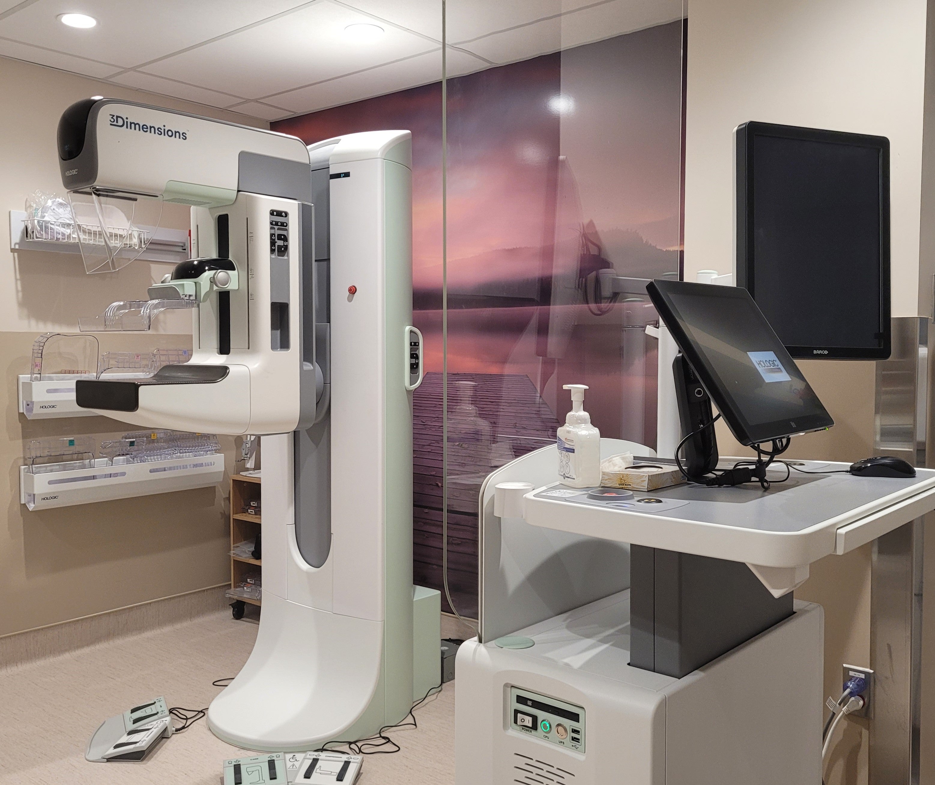 A tall scanner for mammograms in a medical facility stands next to a picture of a sunset and computer equipment.