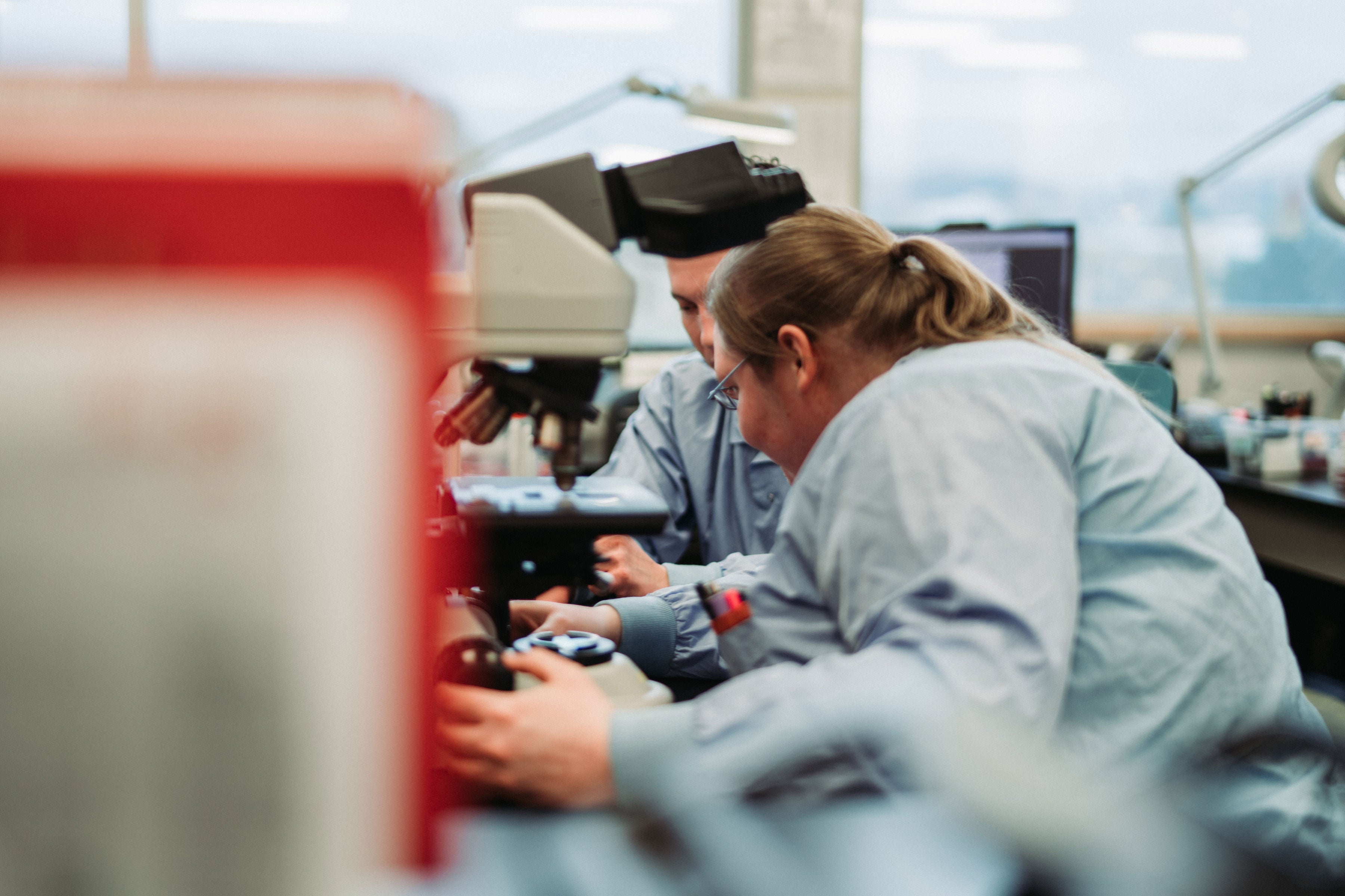 Two people wearing medical scrubs examine something under a microscope.