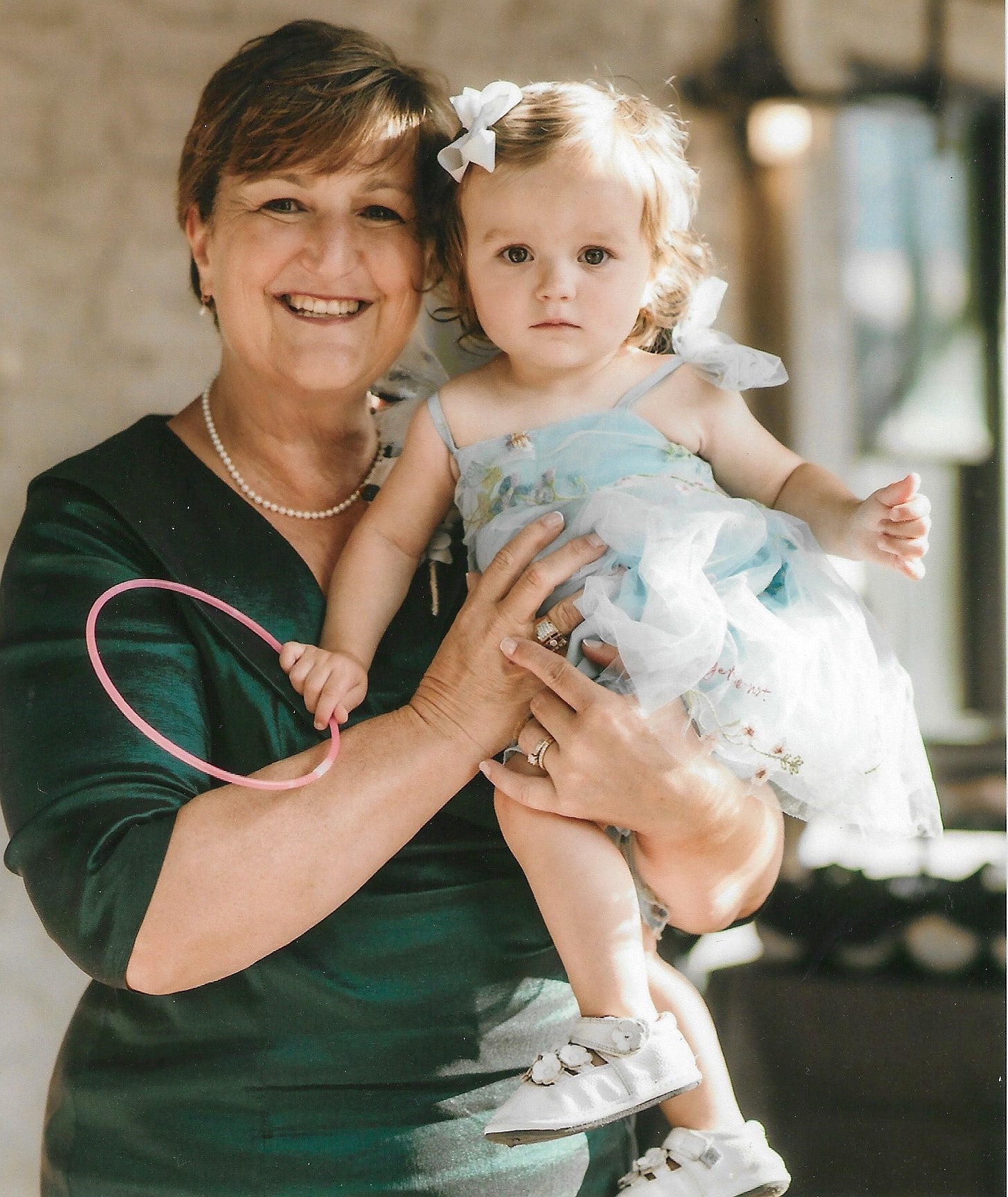 A person with short brown hair wearing a green formal dress holds a young child holding a hoop, wearing a light blue dress and white sandals.