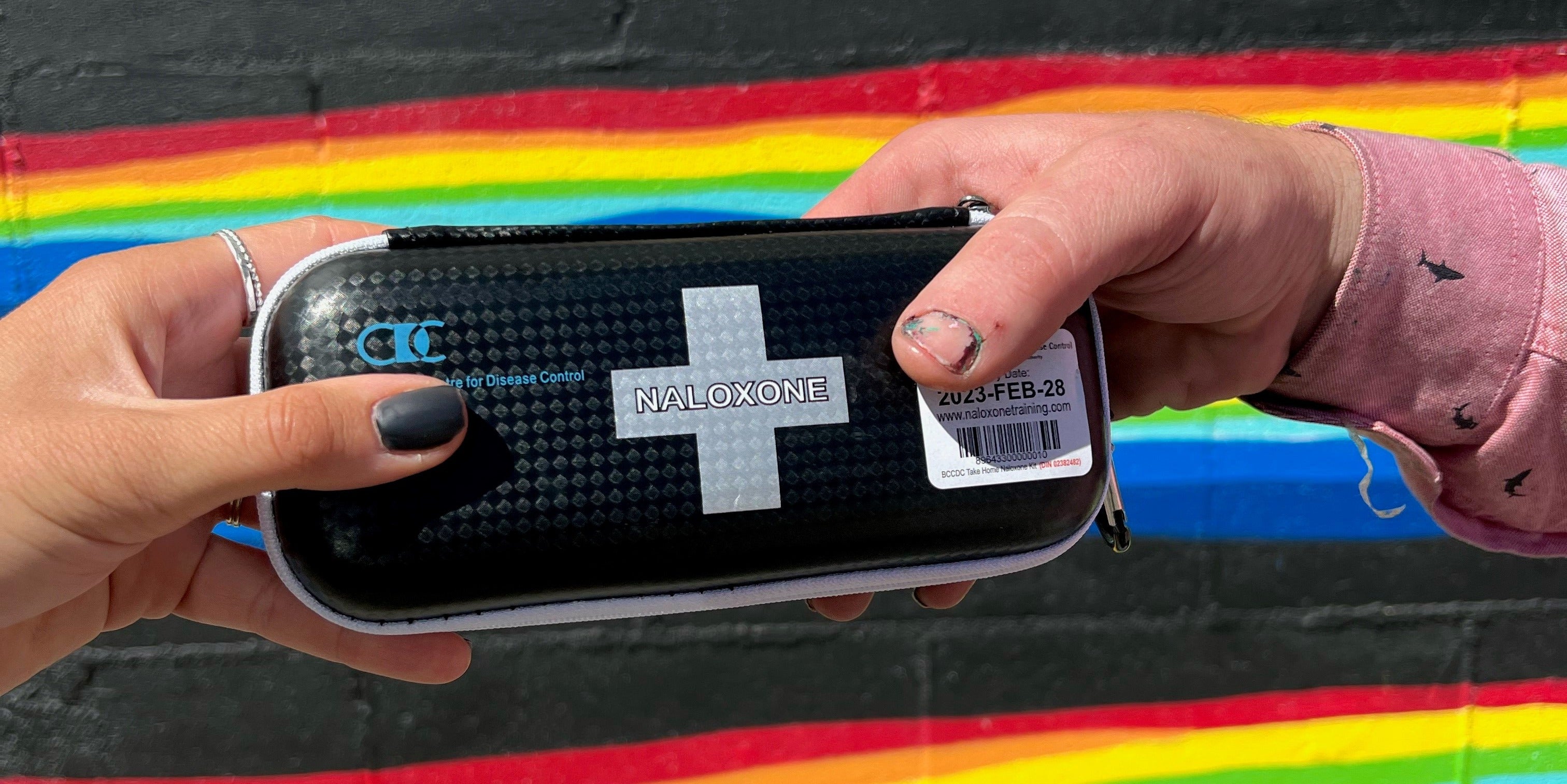 Two hands hold a black case with a grey cross in the middle that says naloxone. There's a rainbow mural in the background.