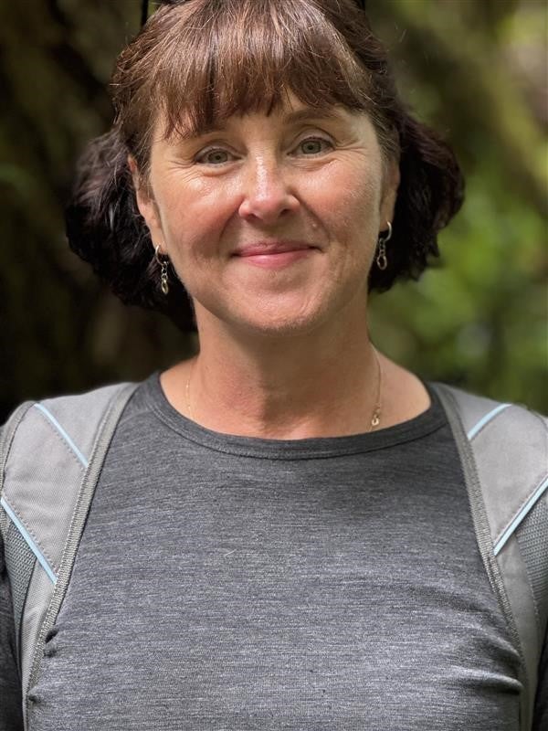 A smiling person with bangs and their hair tied back, wearing a grey cotton shirt.