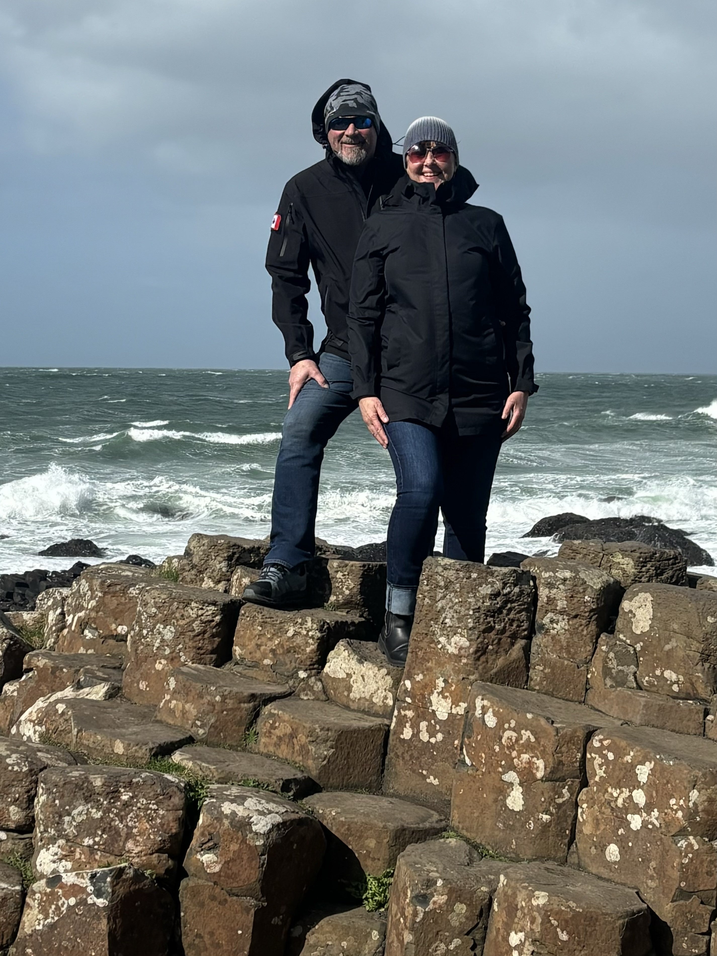 Two people in warm jackets and toques stand on rugged rocks in front of a frothy ocean.