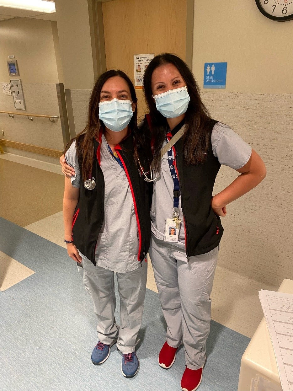 Two women stand side by side with their arms around each other's shoulders in a hospital hallway. Both are wearing grey uniforms, black vests, face masks and stethoscopes