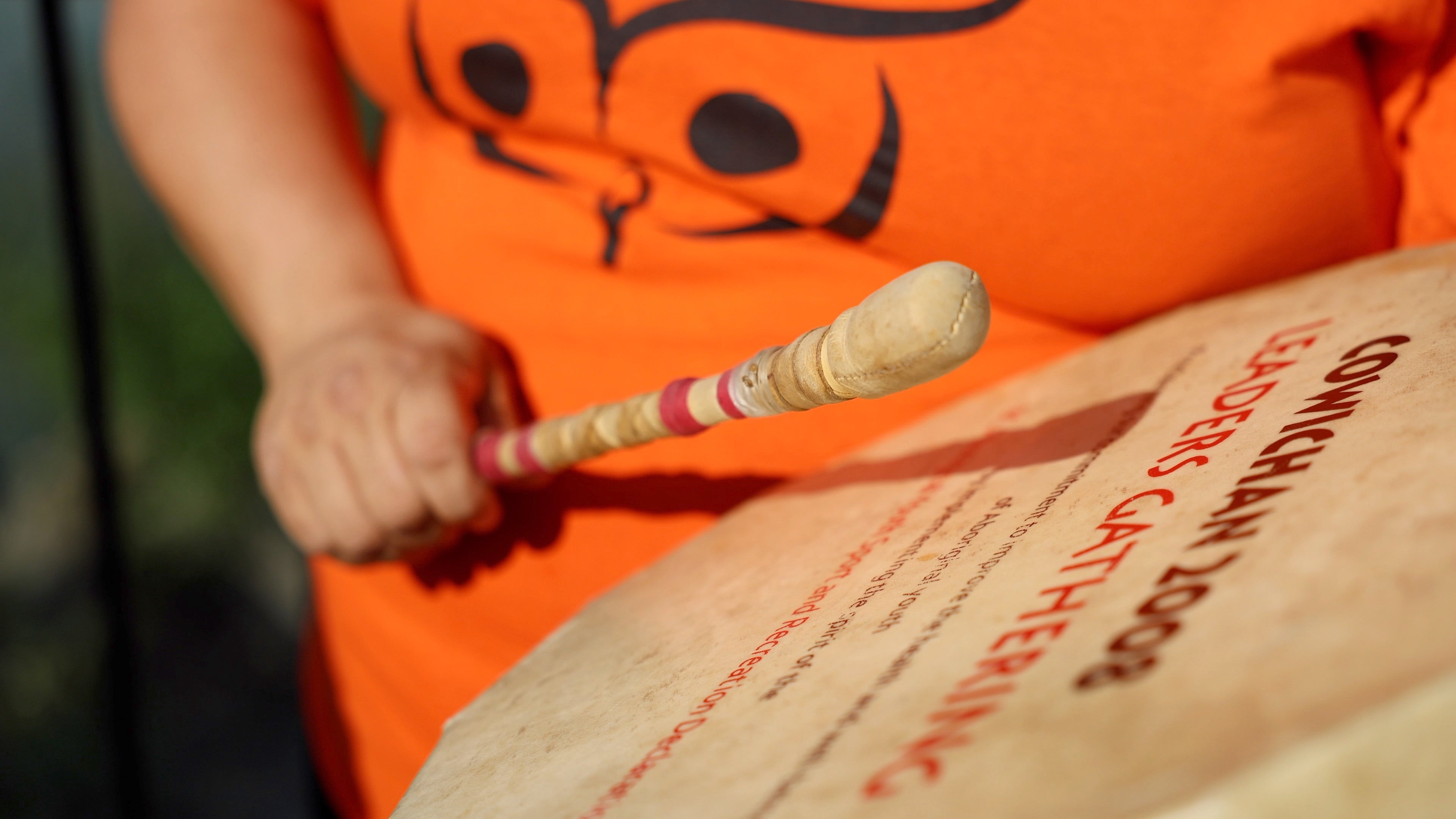 A close up of a person drumming. The drum lettering says Cowichan 2008. They're wearing an orange shirt with an Indigenous motif.