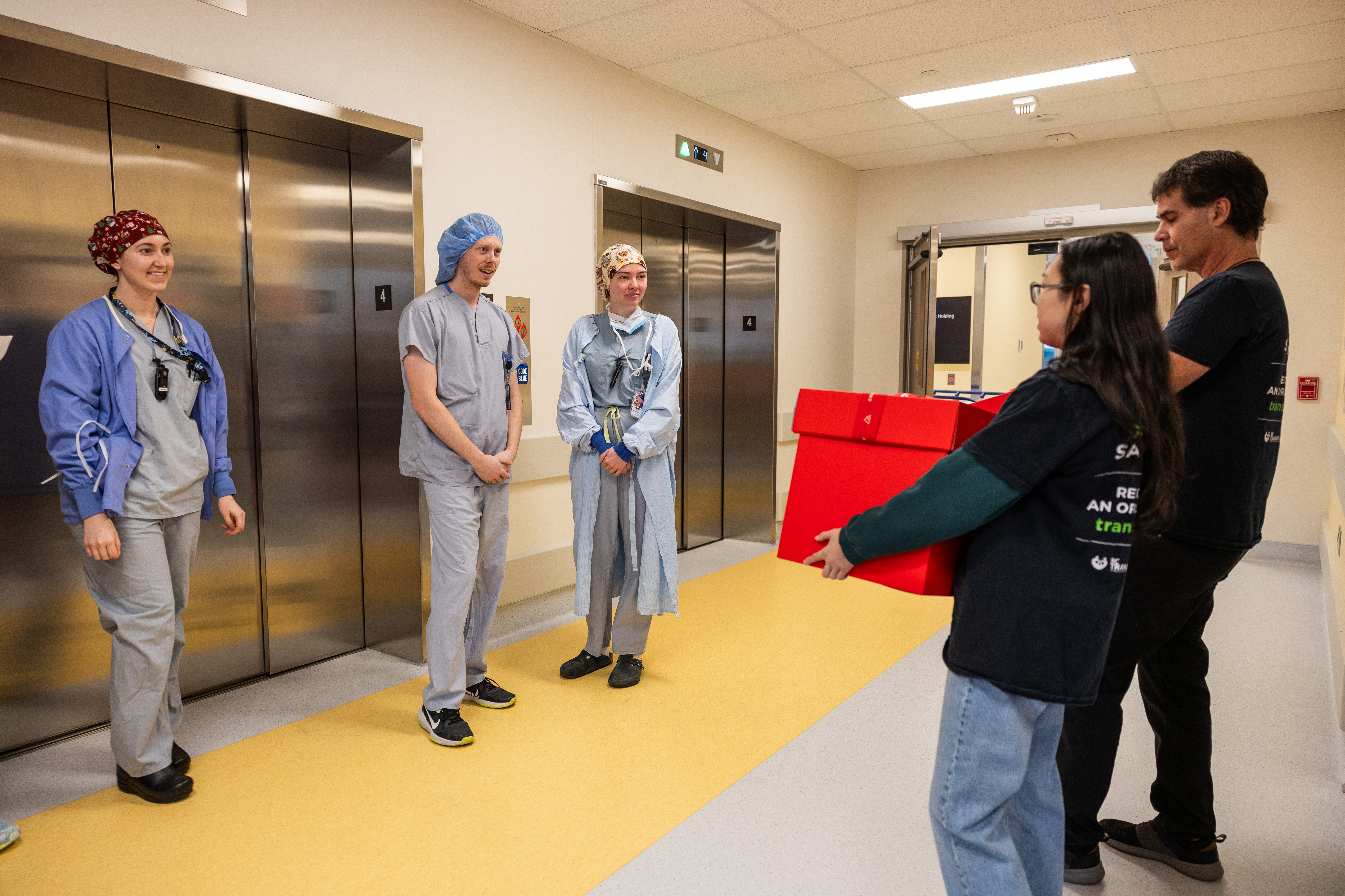 Three people in medical garments stand in front of a bank of elevators while two people carrying big red boxes look on.