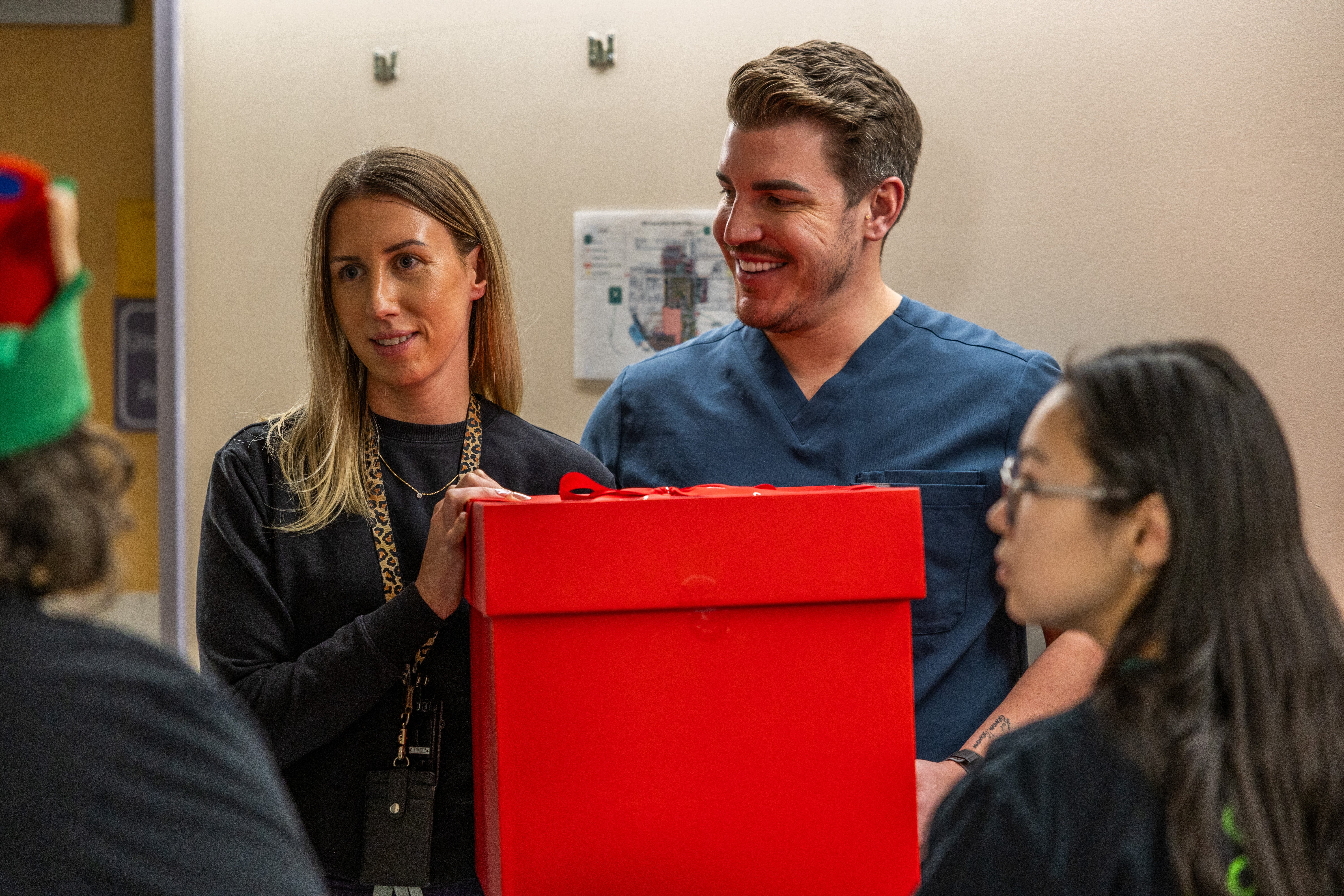 Three smiling people stand together in a hallway. One is holding a big red box.