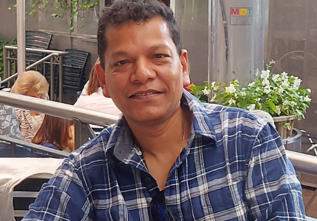 A person wearing a blue plaid shirt with sleeves rolled up leans on a table on an outdoor patio.