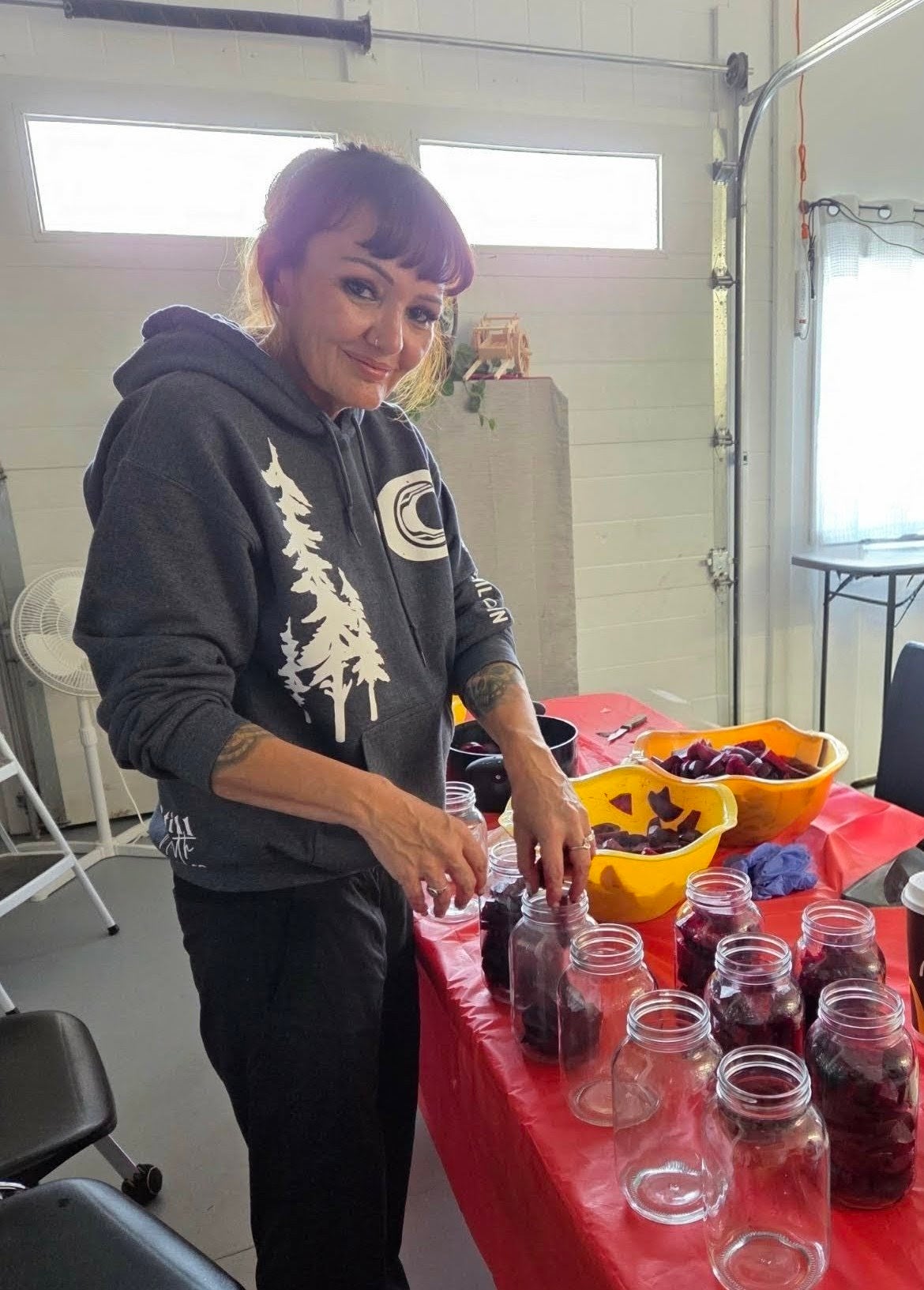 A person wearing a grey hoodie and hair tied back prepares beets for canning at a table covered in a red tablecloth.