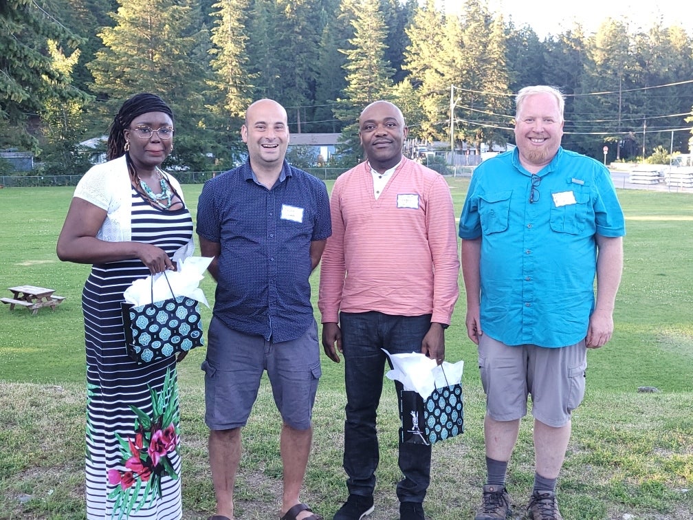 Four people stand in the grass at a park, two of them holding presents.