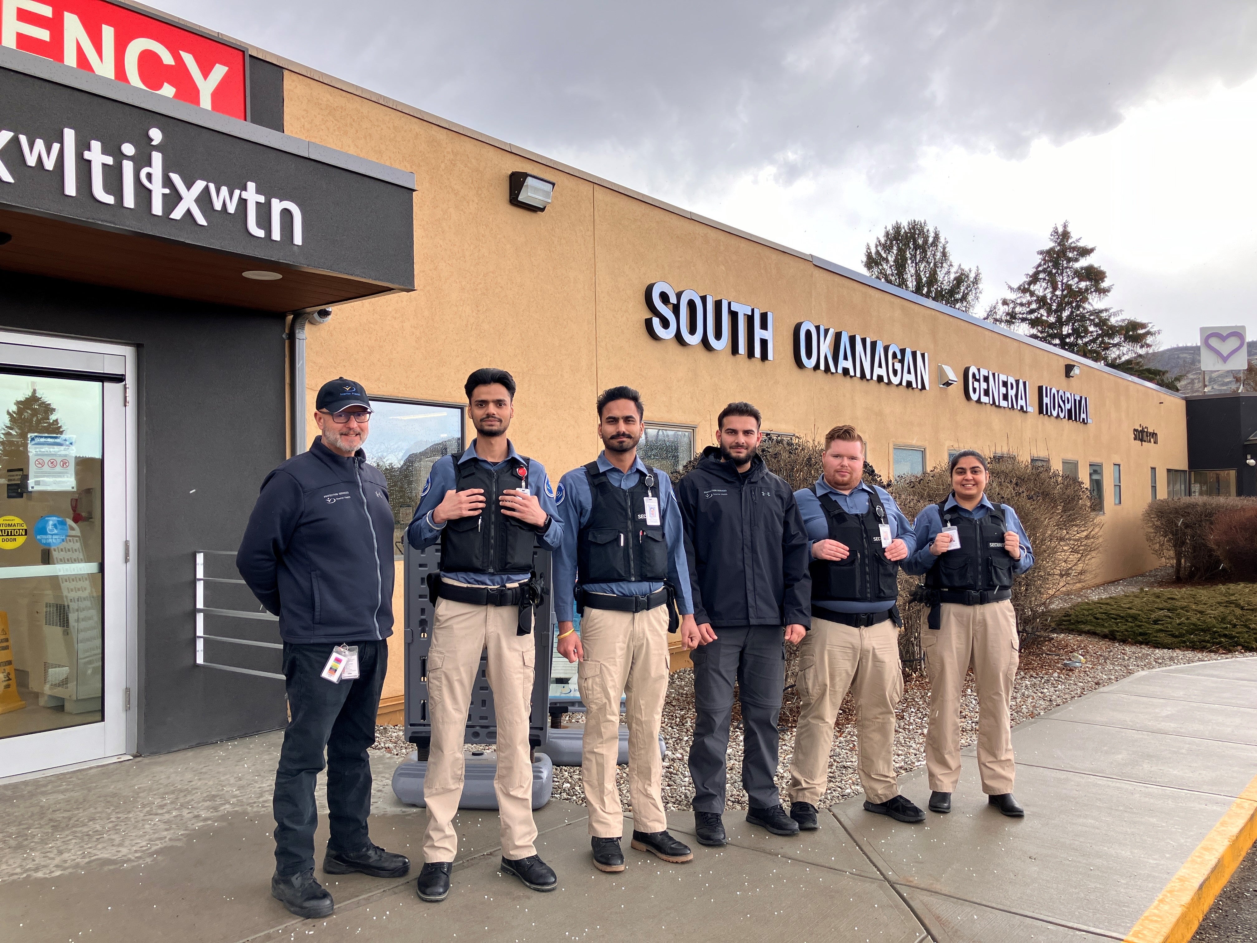 A group of six diverse men stand in security uniforms in front of a beige and grey building with the words South Okanagan General Hospital.