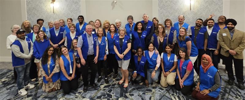A large group of people all wearing blue vests stand and kneel for a photo in a hotel ballroom.
