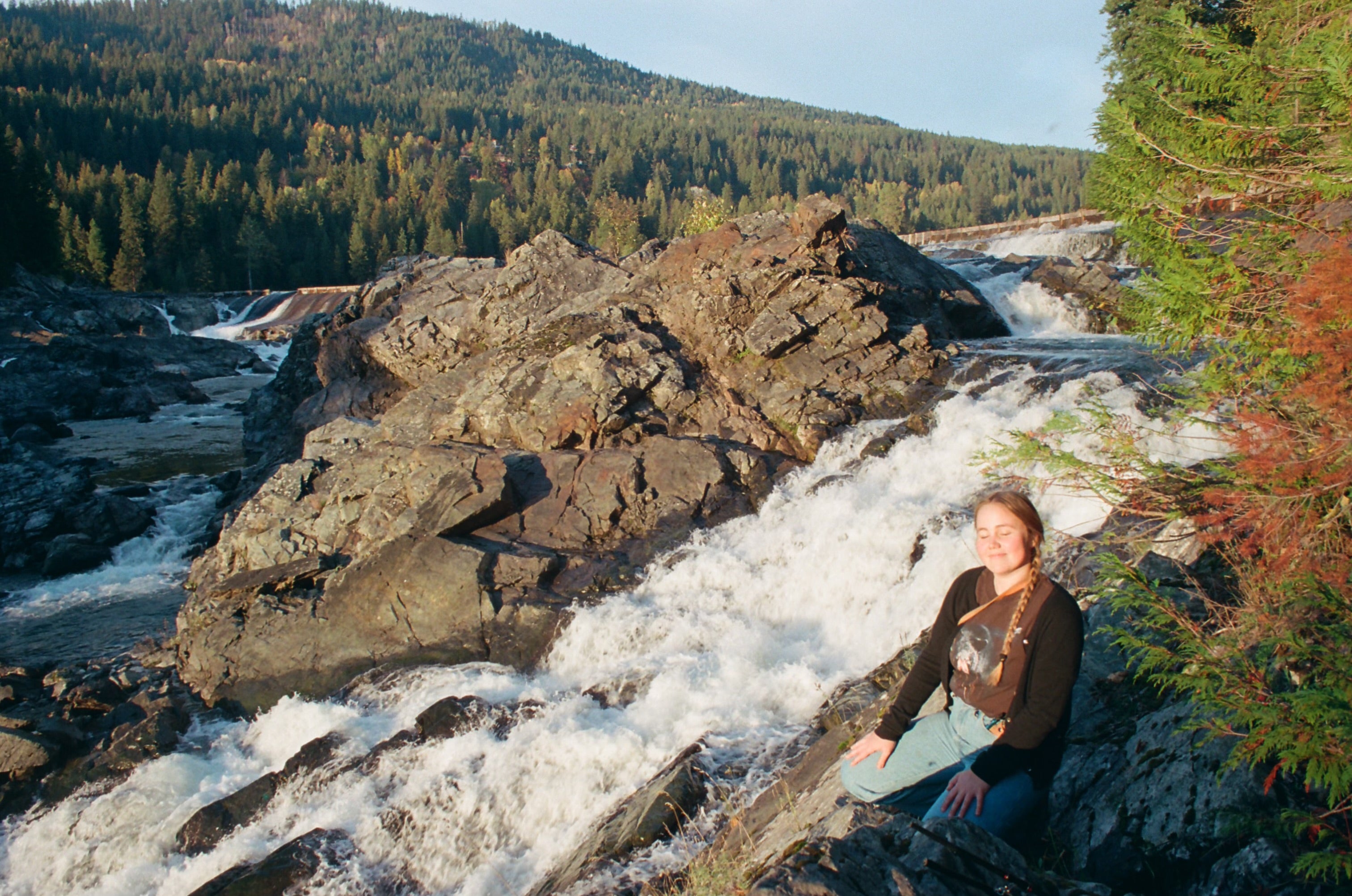 Person sitting on rocks beside fast-moving river rapids in a forested landscape.