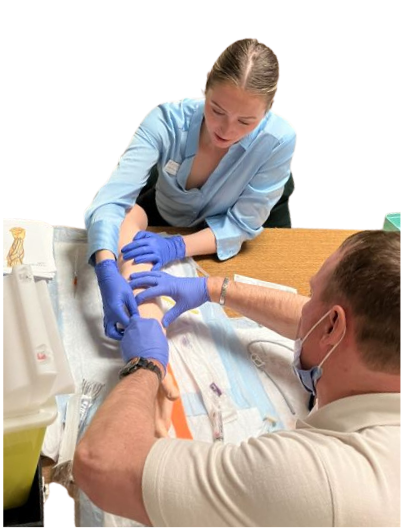 Two medical professionals wearing blue latex gloves practise a procedure on a mannequin arm on a table with a sharps container and gauze pads.