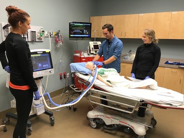 Three people wearing blue medical gloves stand in a hospital room with a simulated patient on a guerney.