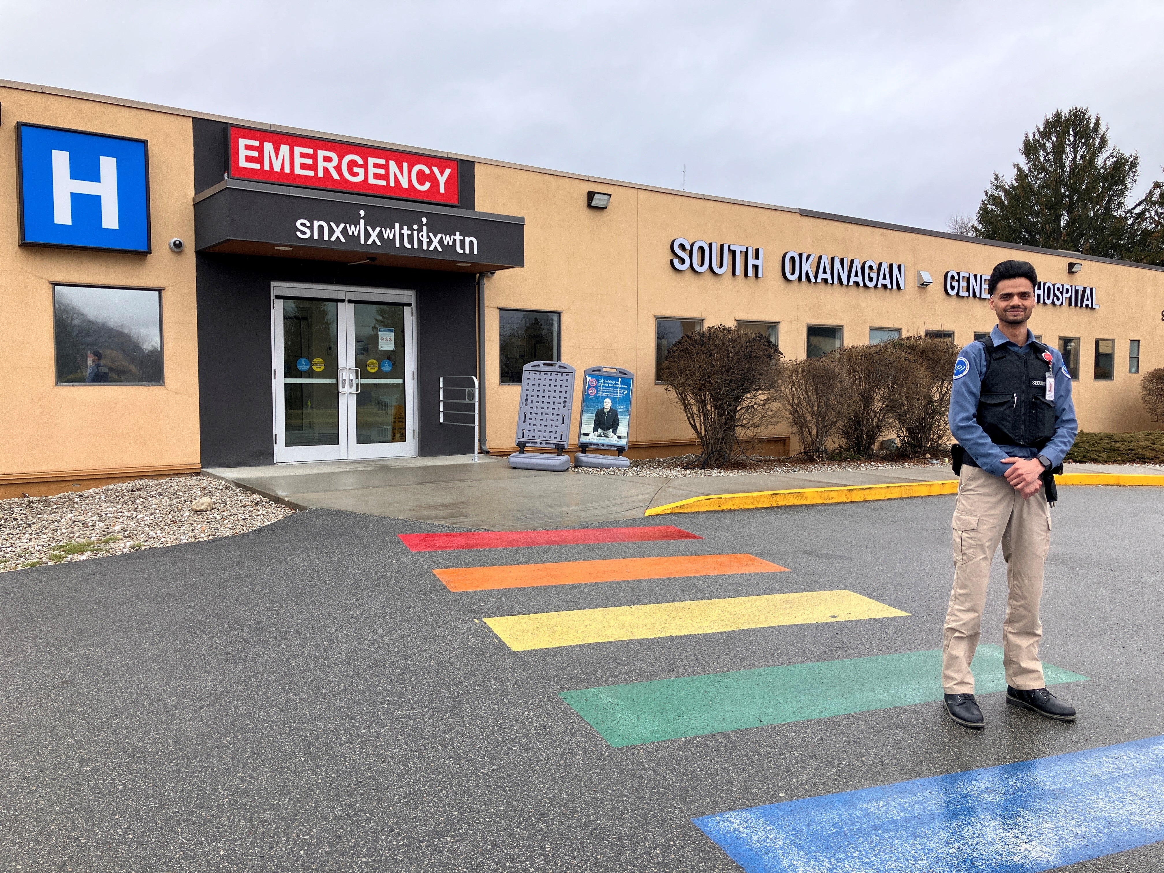 A man with dark hair and a beard stands in a security uniform with his arms crossed in front of a beige building that says emergency, Indigenous lettering and South Okanagan General Hospital.