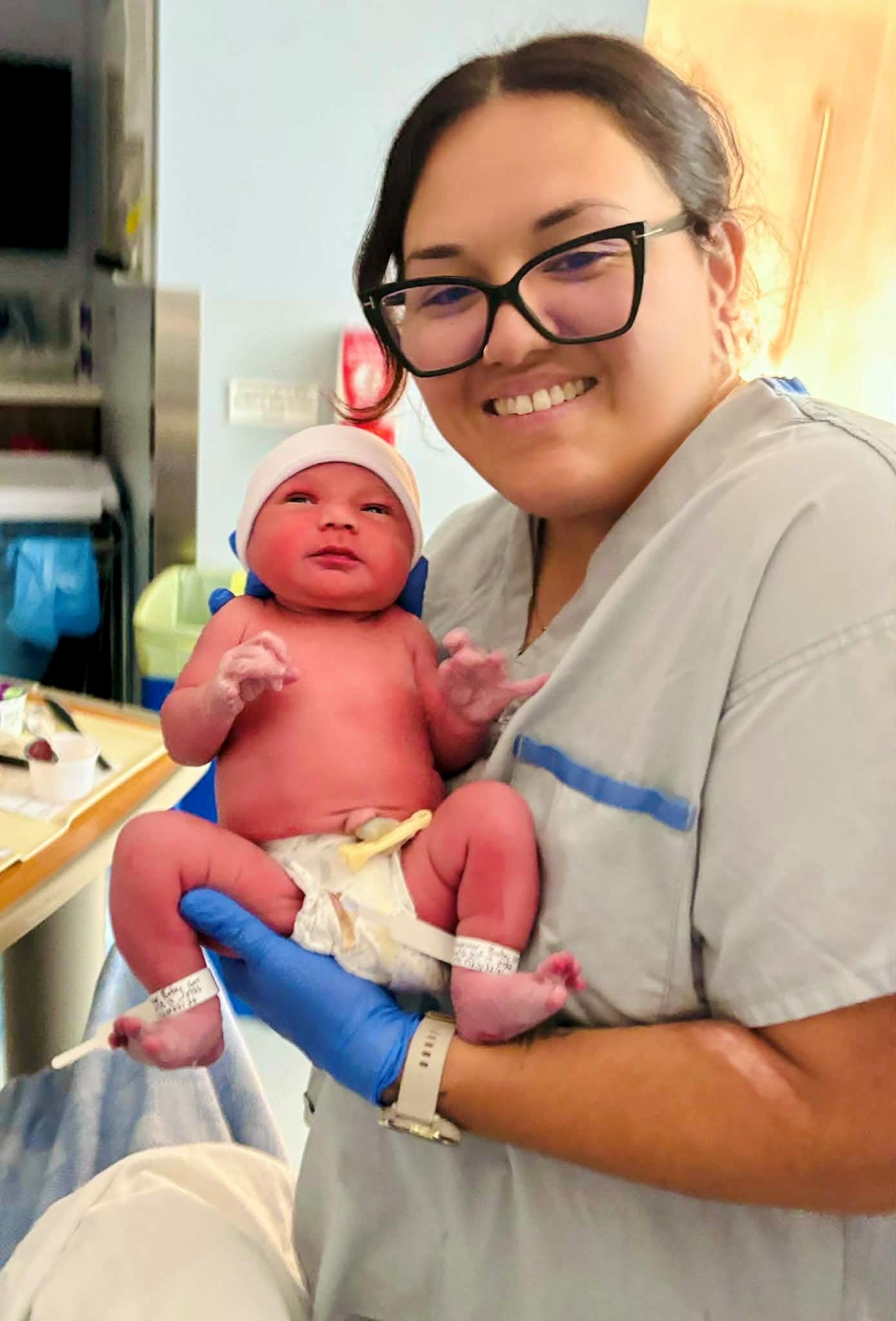 A person with big black glasses wearing a medical coat and blue gloves holds a newborn baby.