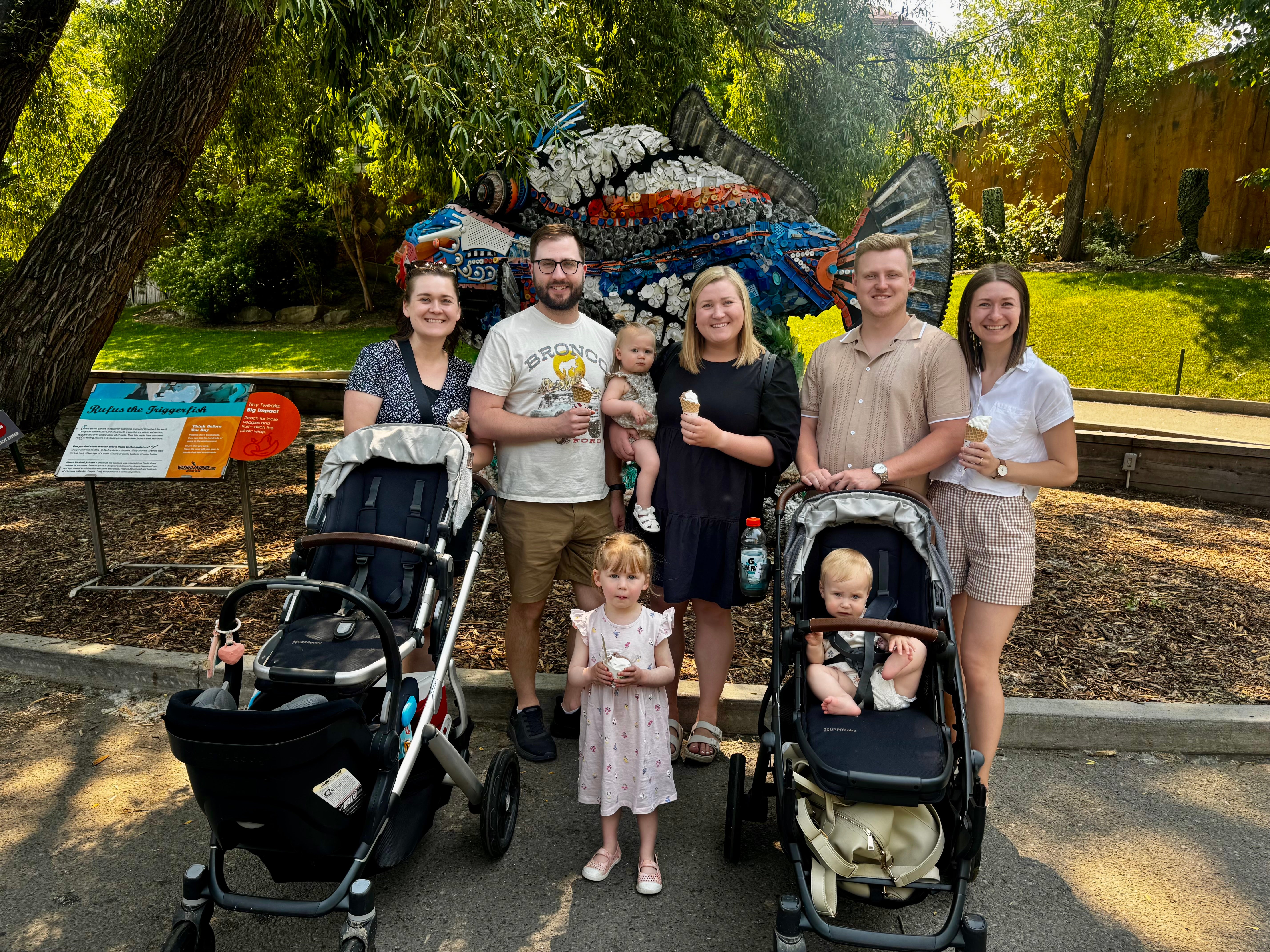 Family, including a baby and two toddlers, and five adults, pose and smile in front of a statue of a fish. Some hold ice cream cones.