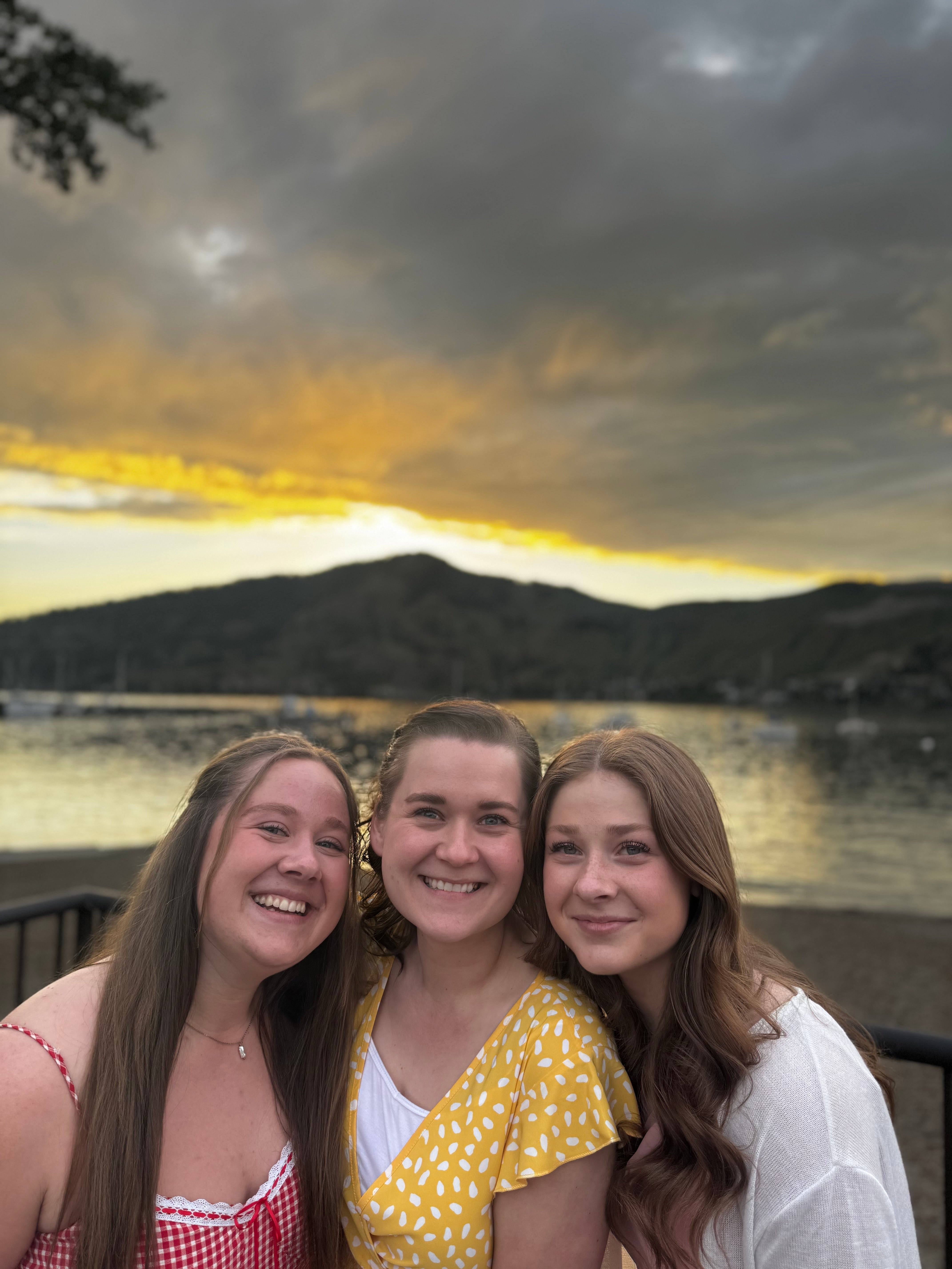 Three people have their arms around each other, as they stand together smiling in front of a lake at sunset. There are mountains in the background. 