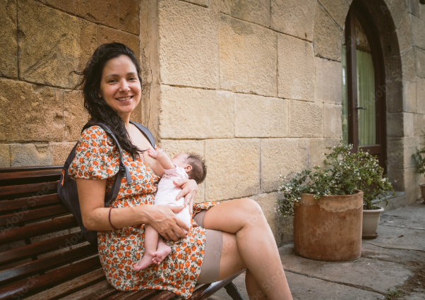 A person in a short brown flowered dress nurses a baby on a bench next to a building.
