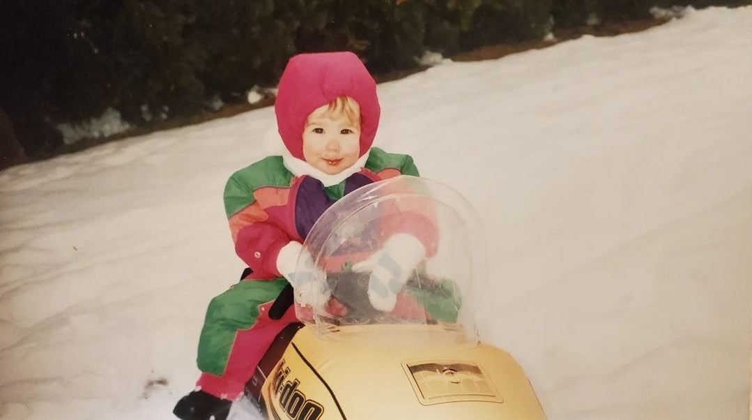 A child in colourful winter clothing sits on a yellow snowmobile in the snow.