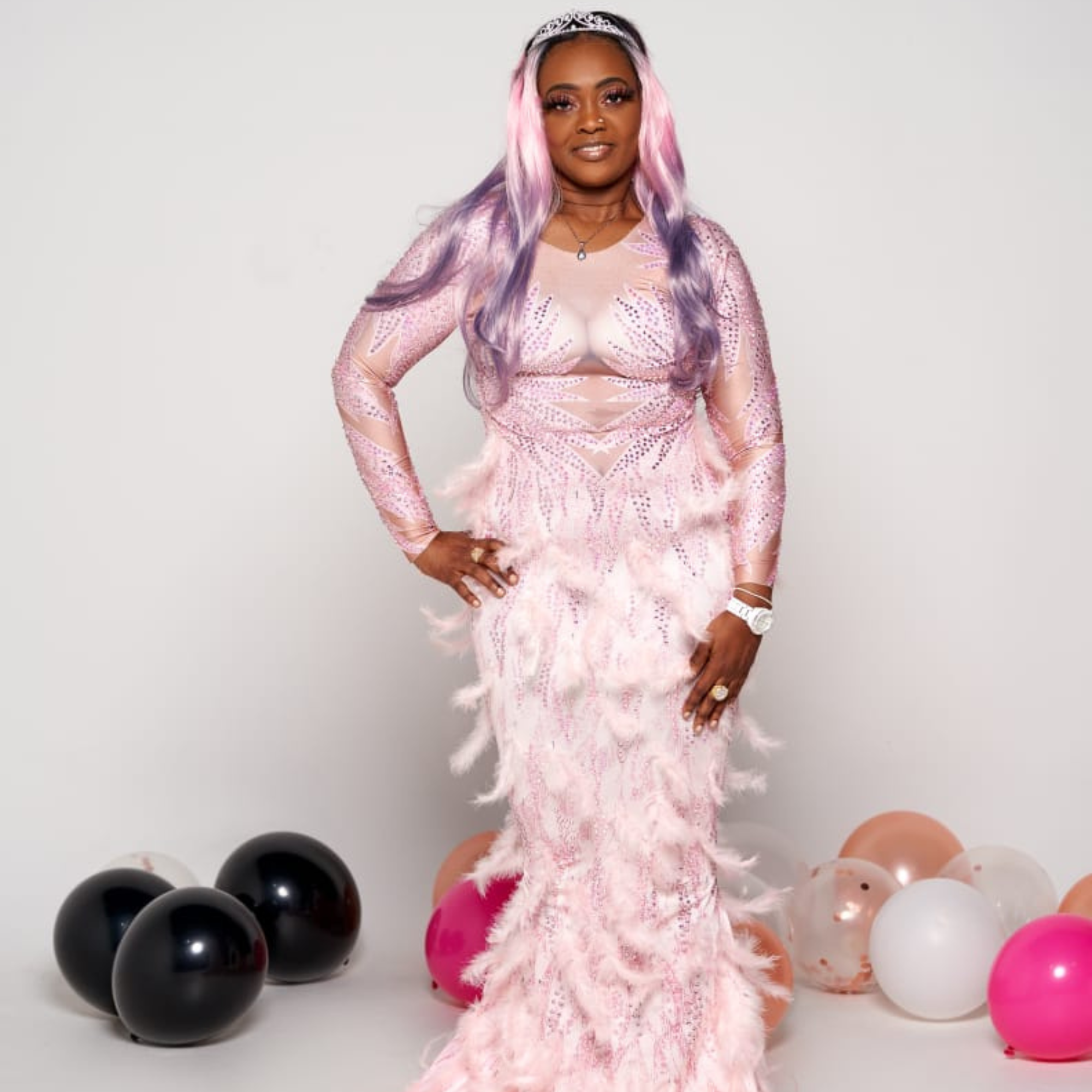 A smiling woman with blonde, pink and purple hair wearing a pink feathered floor length gown standing in front of balloons that are laying on the floor.