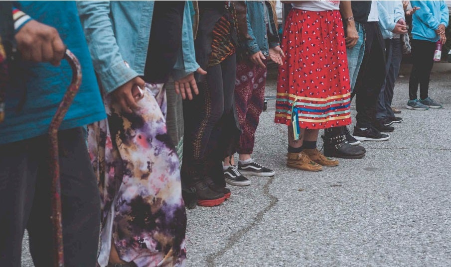 Foot and leg shot of a group of people standing side-by-side in a line, appearing to be Indigenous and in prayer.