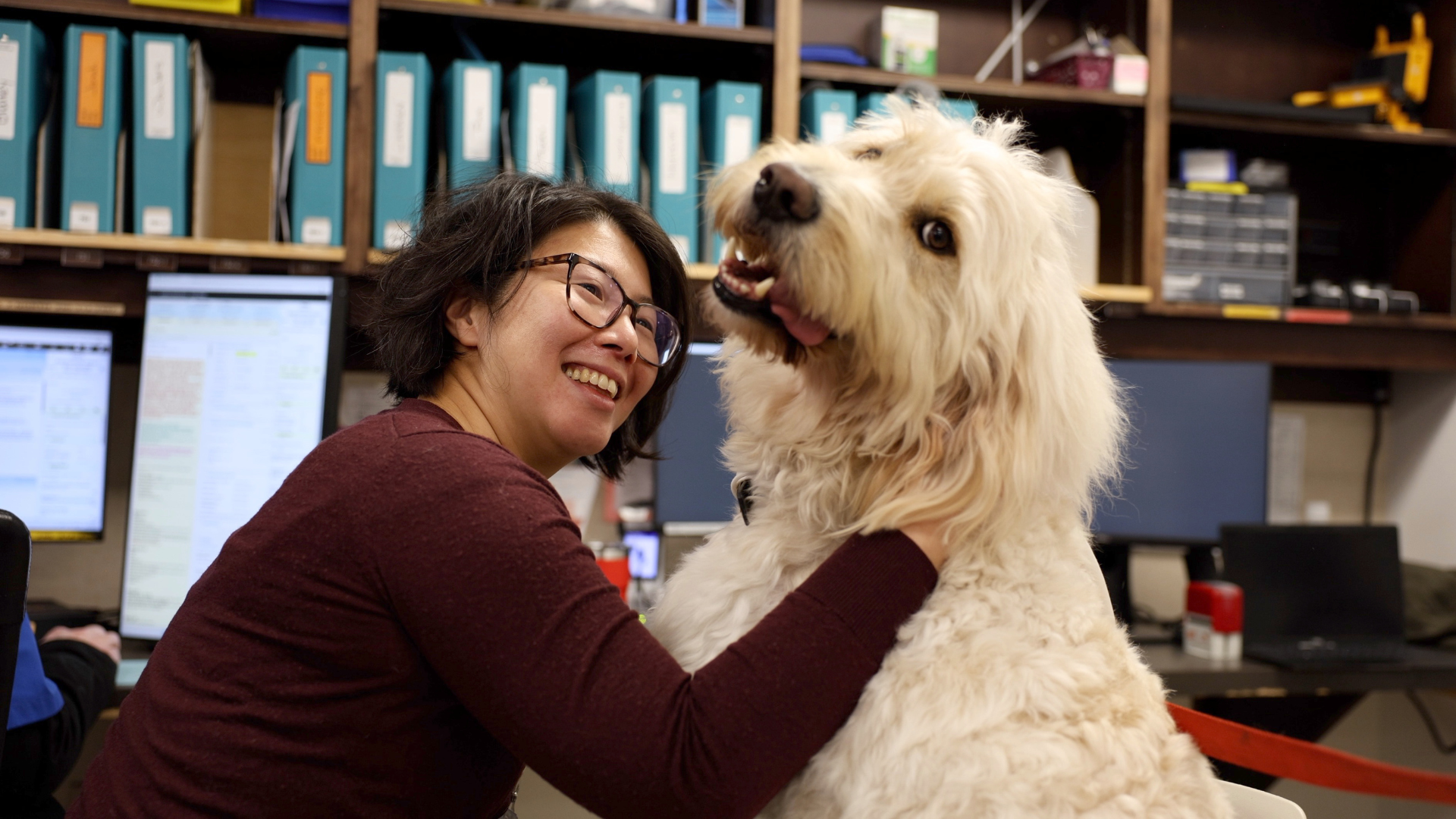 A health-care staff member in a red shirt hugs a white fluffy dog at a hospital.