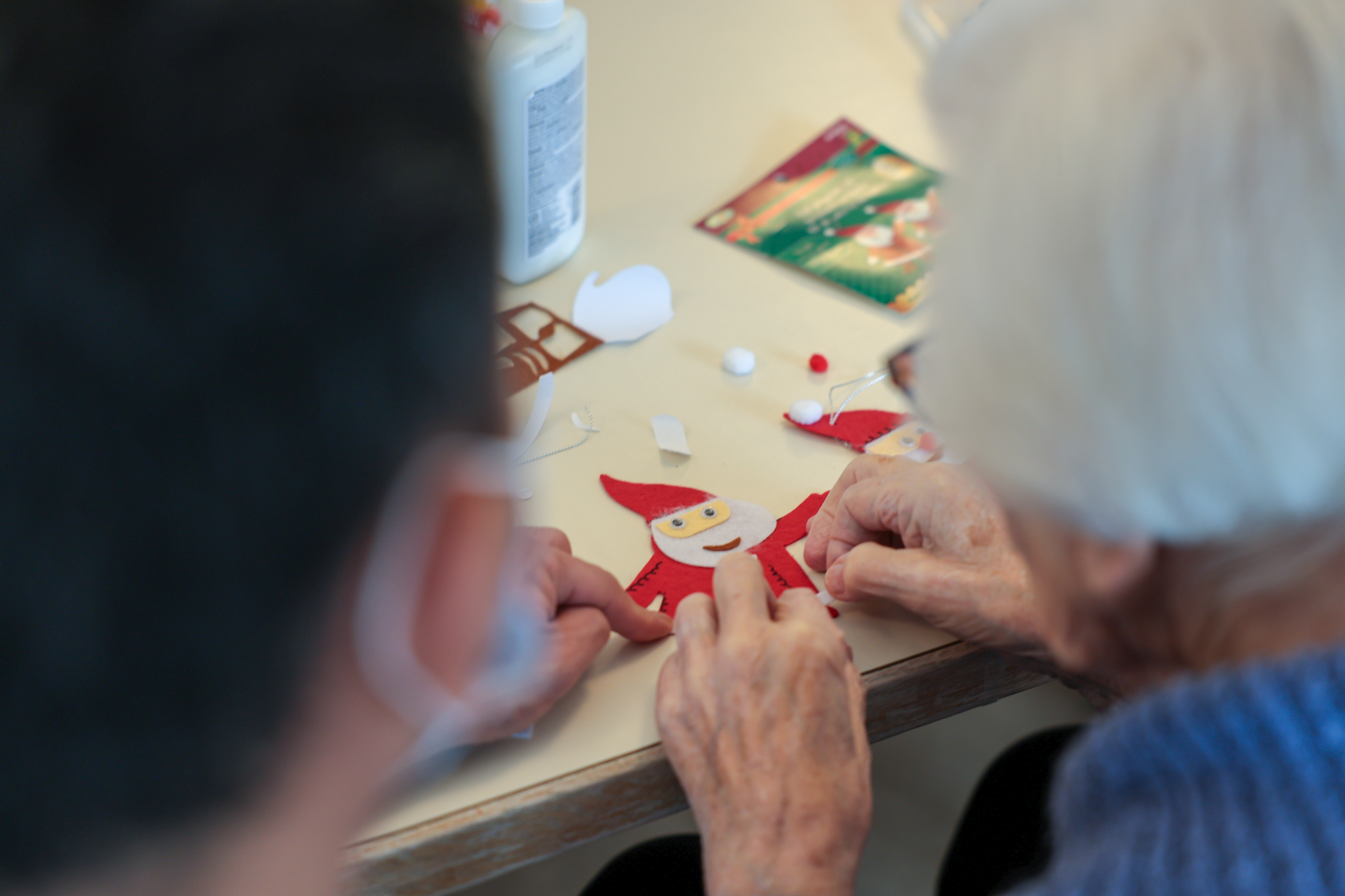 An elderly person with white hair works on a Christmas craft sitting next to a person with dark hair