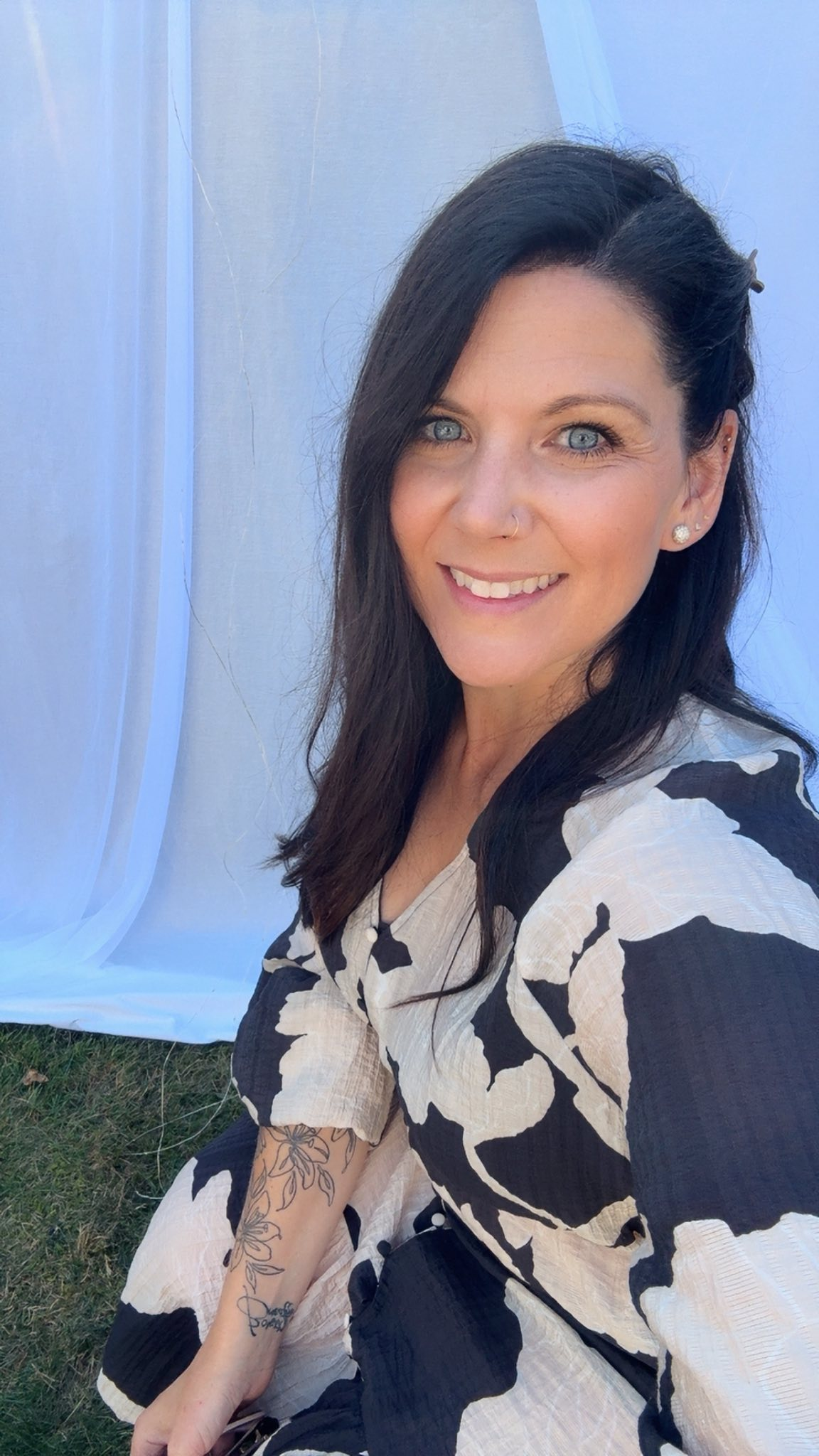 A person in a black and white blouse with long black hair sits on the grass in front of a tent.