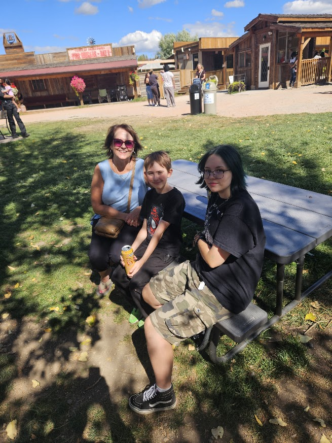 An adult wearing sunglasses and two children pose for a picture on an outdoor bench with some old time wooden buildings in the background.