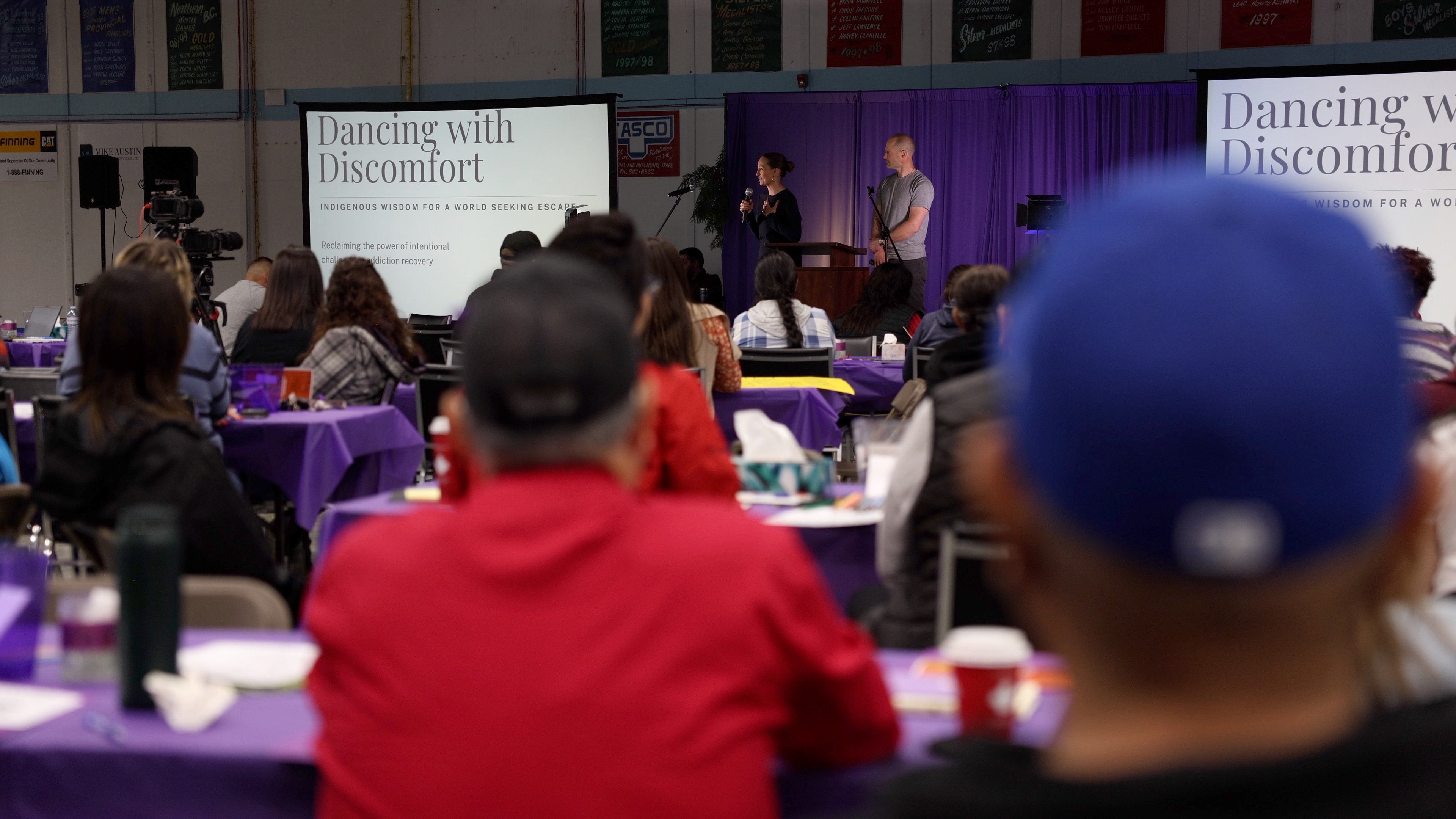 People sitting at round tables pay attention to two speakers at a podium. There are presentation screens that read dancing with discomfort.