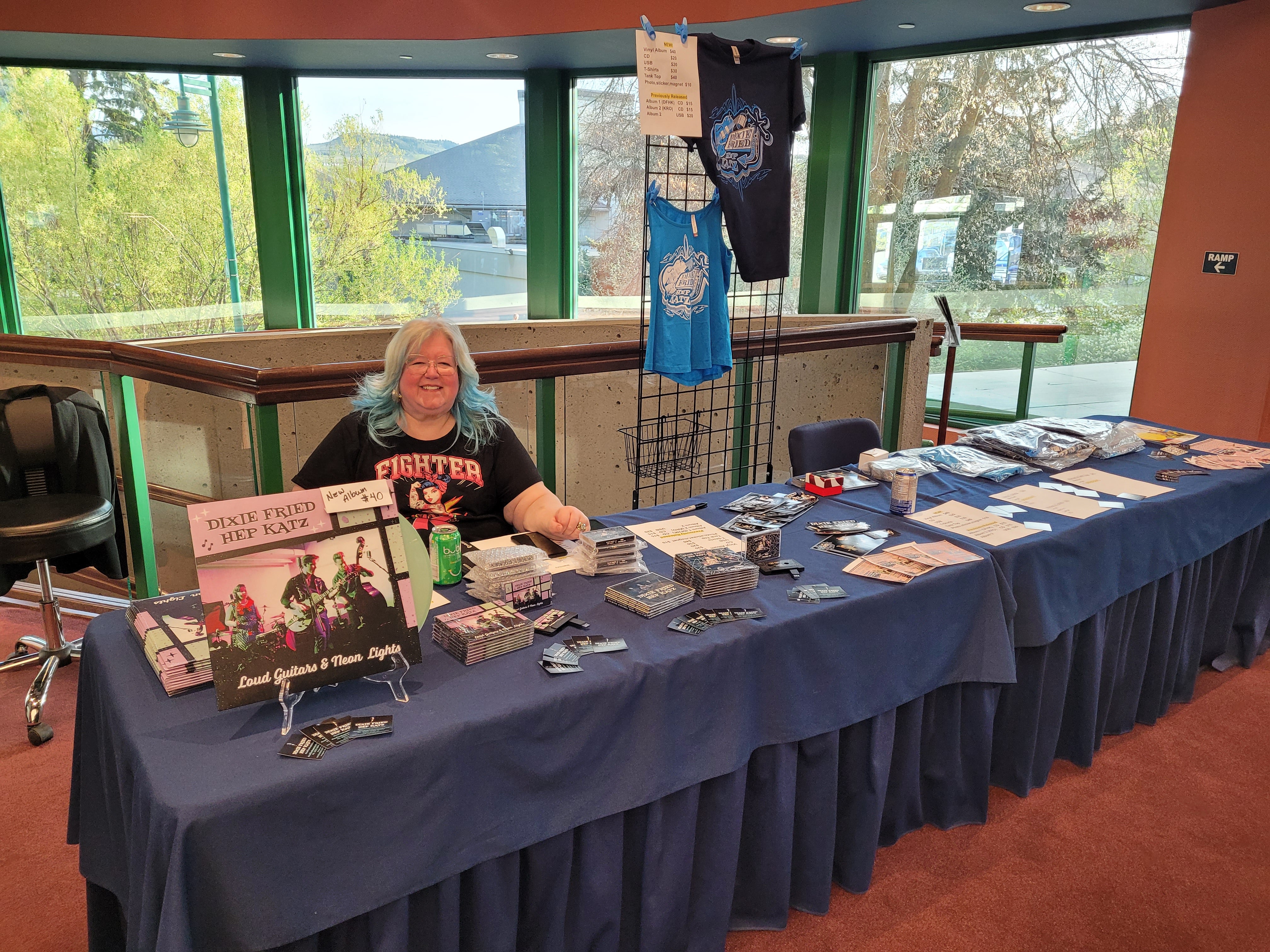 A person is sitting behind a band merch table smiling. They are wearing a black graphic tee shirt and have white, blonde hair that is dyed blue on the ends.