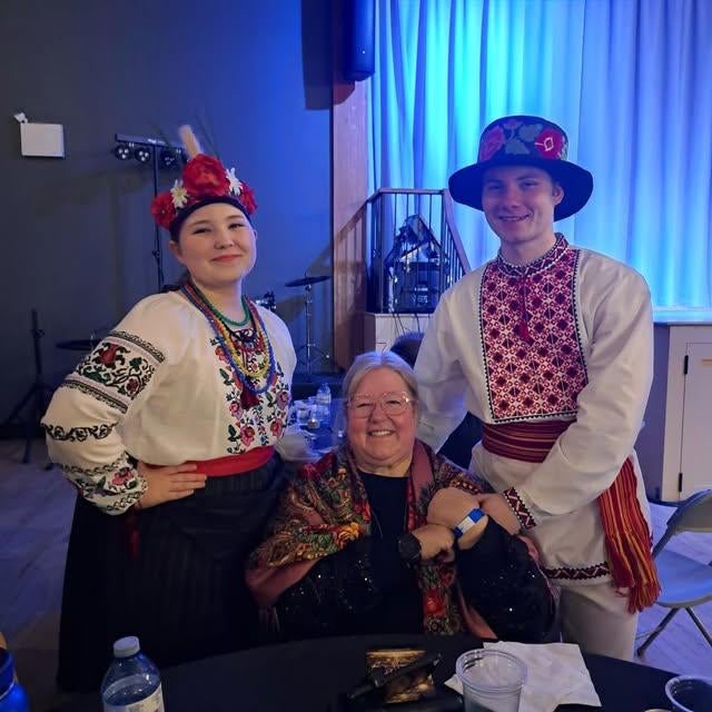 A person is sitting in a chair, with their niece and nephew standing on either side of them. They are dressed in Ukranian dance costumes and are smiling for the photo.