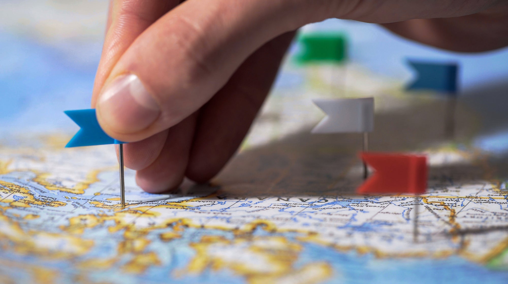 A hand puts a blue pin on a laid out map that already has four other pins on it (red, white, green and blue).