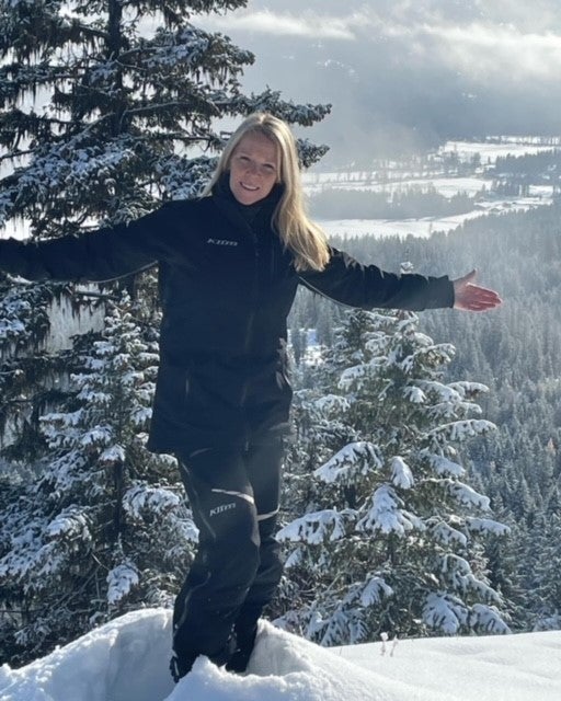 Long-haired blonde women wearing black ski jacket and ski pants with arms outstretched, stands in snow with snow-covered trees and mountains in background.