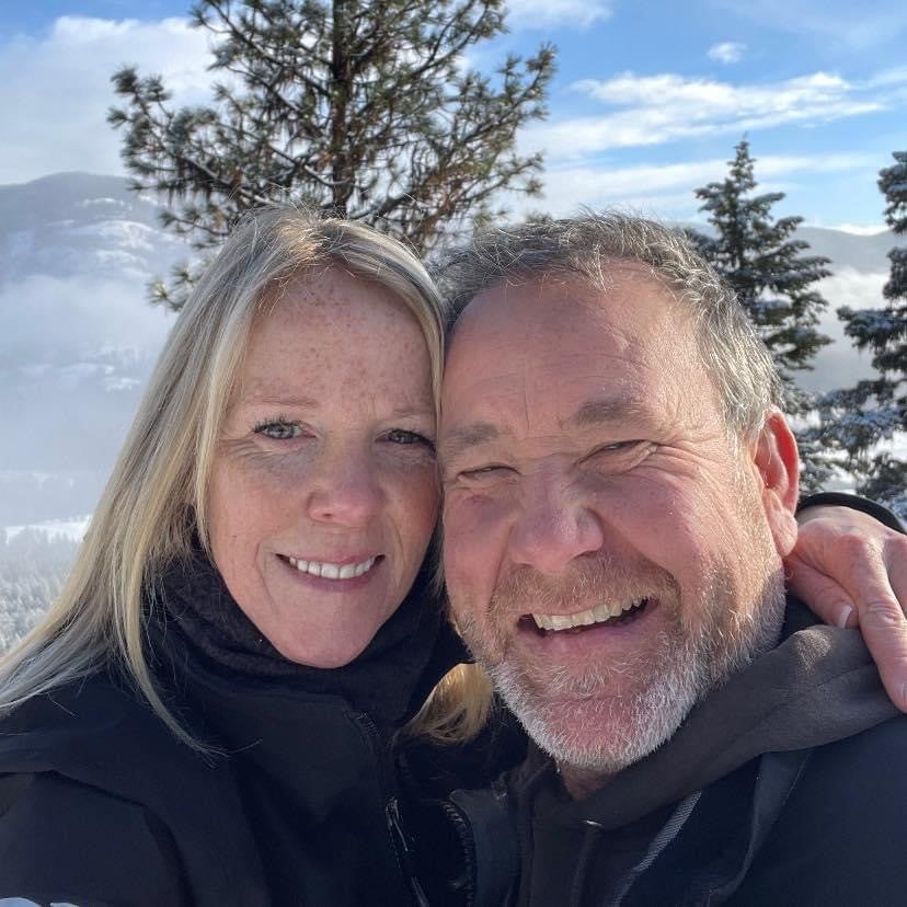 Blonde woman with arm around man with grey beard, wear black winter jackets, with trees, snow, and clouds in the background.