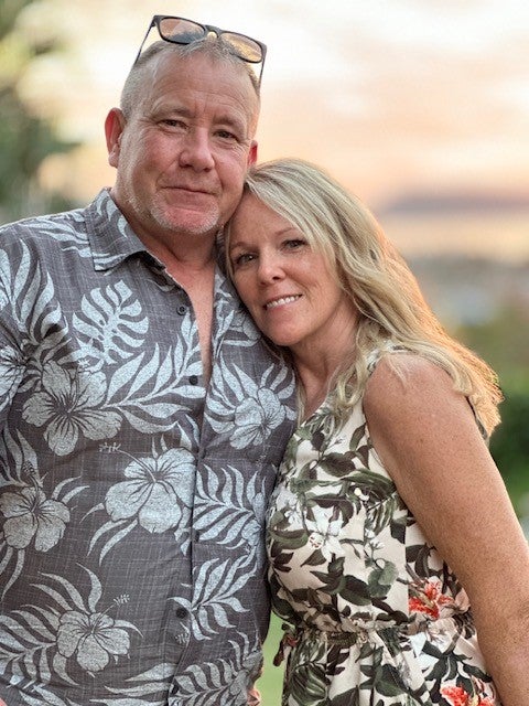 Middle-aged man wearing grey buttoned shirt with white leaf and flower pattern and sunglasses on head stands with long-haired blonde woman resting her head on his shoulder.